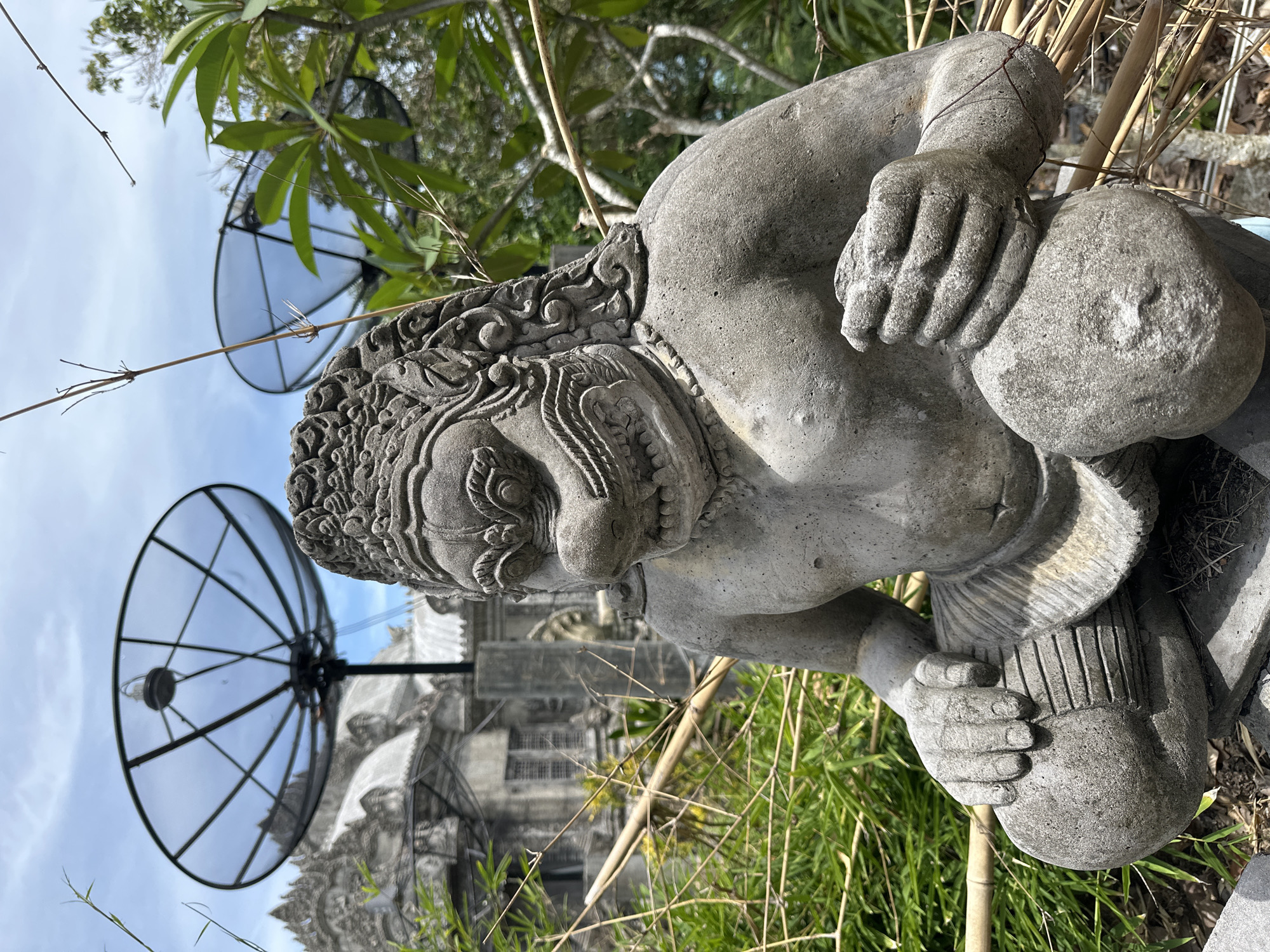 The image shows a stone statue of a fearsome creature, possibly a demon or ogre, with detailed facial features and ornate headdress. It is seated with its legs bent and its hands clasped around a large, round object. The statue is positioned outdoors amidst lush green foliage and dry bamboo stalks. In the background, two large satellite dishes are visible, mounted against a backdrop of buildings with distinctive architectural elements, suggesting a location in Thailand. The sky is partly cloudy. The overall scene presents a blend of traditional art and modern technology in an outdoor setting. This specific statue is located at the "The Great Buddha" site, also known as Wat Phra Yai, in Ko Pha-ngan District, Thailand.