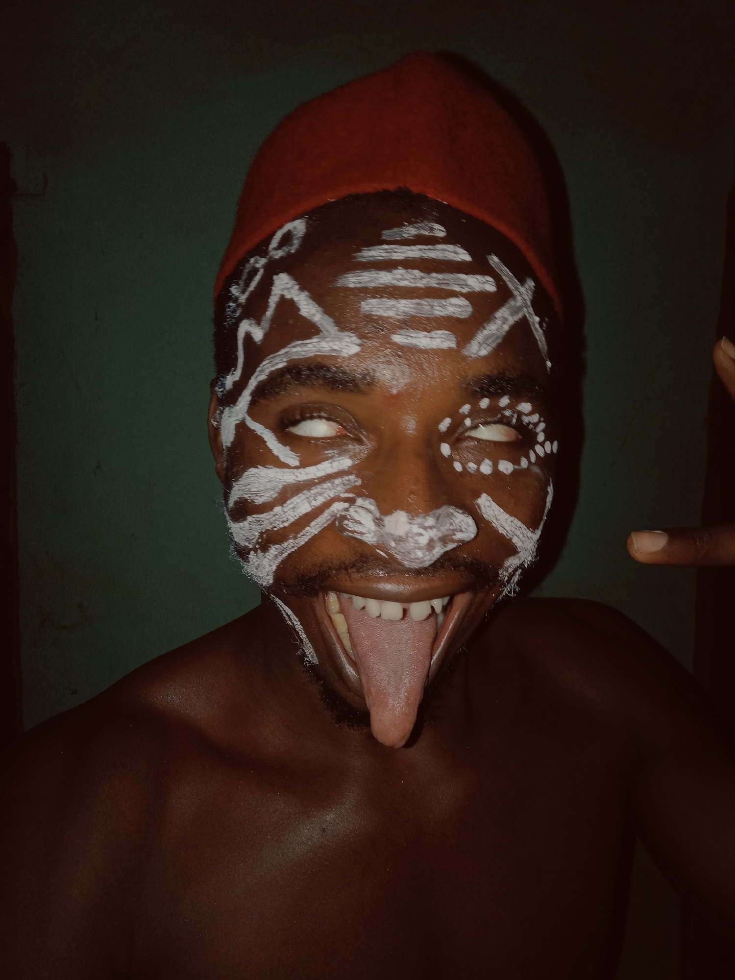 A person with dark skin, wearing a red hat and white paint on their face, is sticking out their tongue. The person's eyes are rolled back, showing only the whites. A finger is visible in the top right corner of the image. The background appears to be a green wall. The location is Unknown, Unknown.