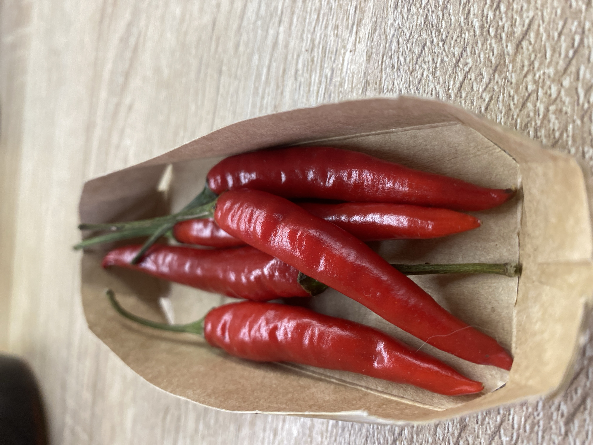 A close-up shows a small, brown paper bag containing approximately six red chili peppers. The peppers are bright red and appear fresh, with green stems visible on some. The bag sits on a surface that looks like light-colored wood with a subtle grain pattern. The image has a slightly blurred background and is taken from a high angle. There is no information about the location in Chaikovsky, Russia.