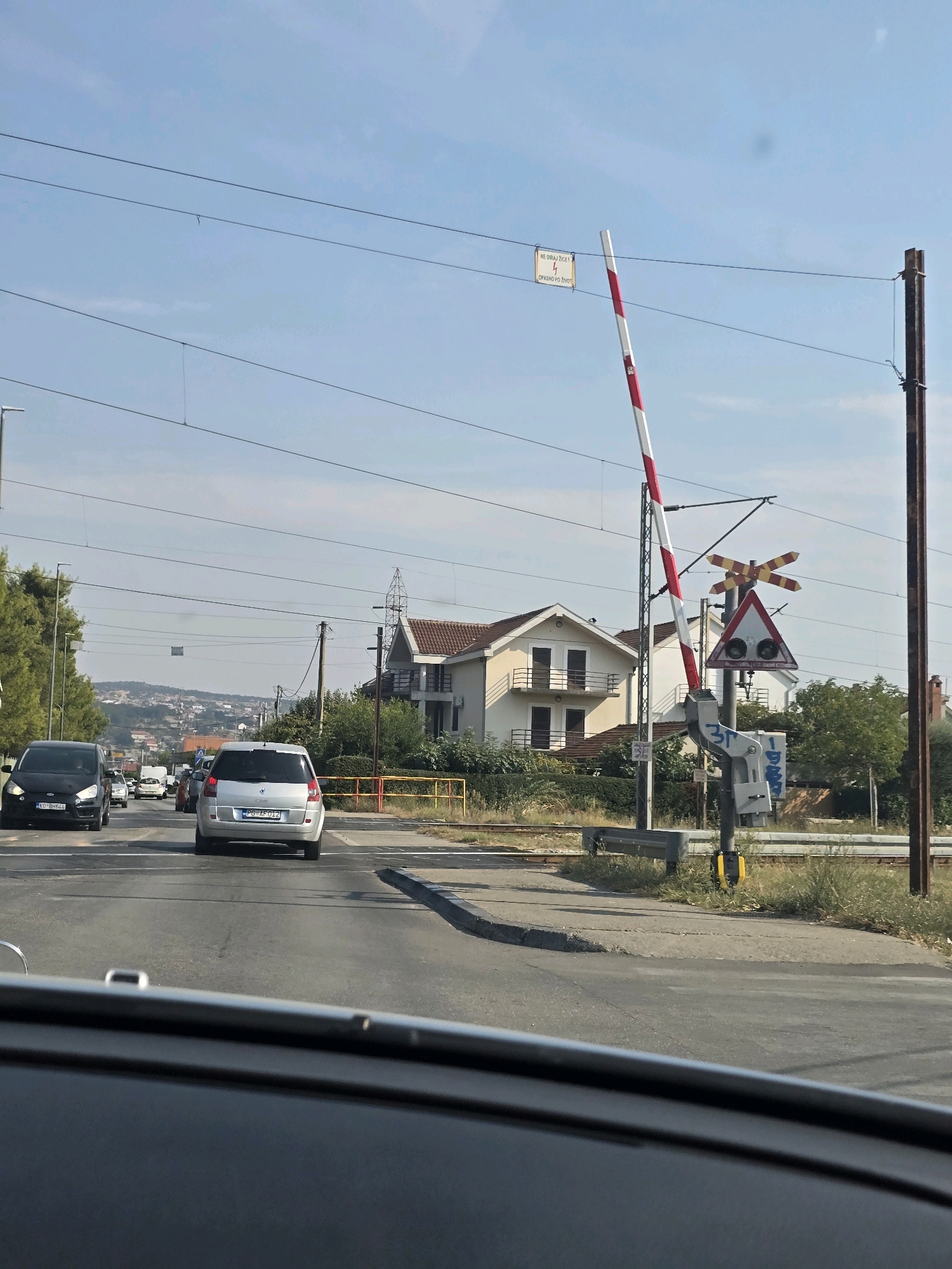 The image shows a view from inside a vehicle looking forward towards a railway crossing in Podgorica, Montenegro. A red and white striped railway barrier is partially lowered. Traffic signs, including a triangular warning sign with two red lights and a crossbuck sign, are visible on a pole. Overhead power lines for the train and possibly street lights are present. Several cars are on the road approaching the crossing. In the background, there are houses and buildings, with a hilly landscape beyond. The sky is clear and blue.