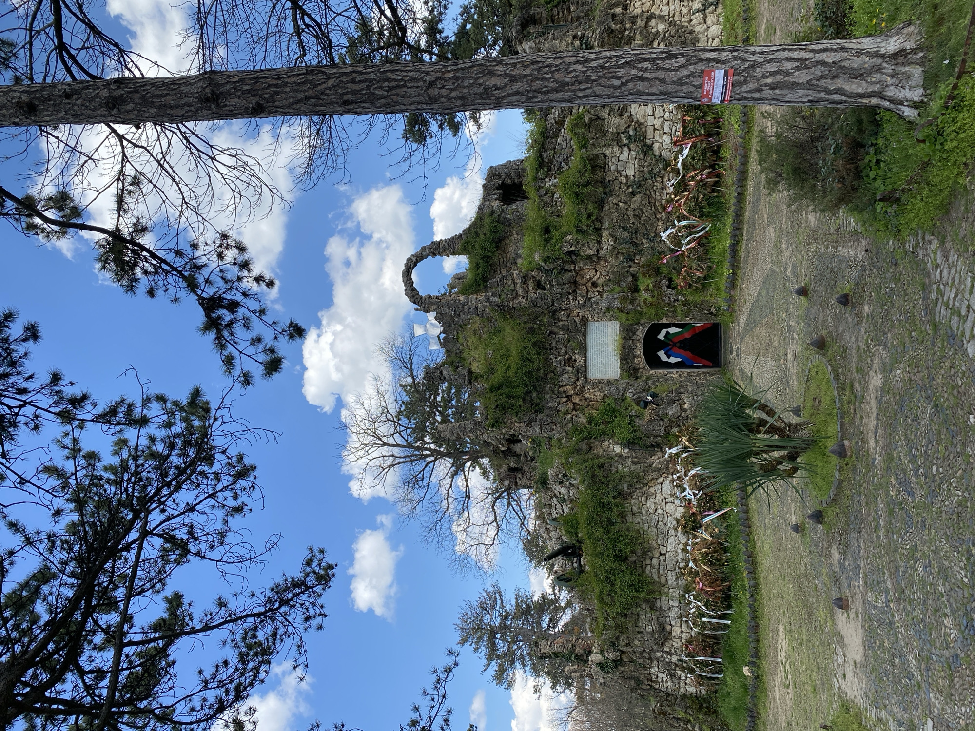 The image depicts a memorial site in Pleven, Bulgaria. A stone structure resembling ruins is visible, with an archway overgrown with vegetation. A plaque is affixed to the structure near an entrance adorned with colors. White ribbons and floral arrangements line the wall. A cobblestone path leads up to the site, with grass patches on either side. Several trees frame the scene, their branches reaching towards a sky dotted with clouds.