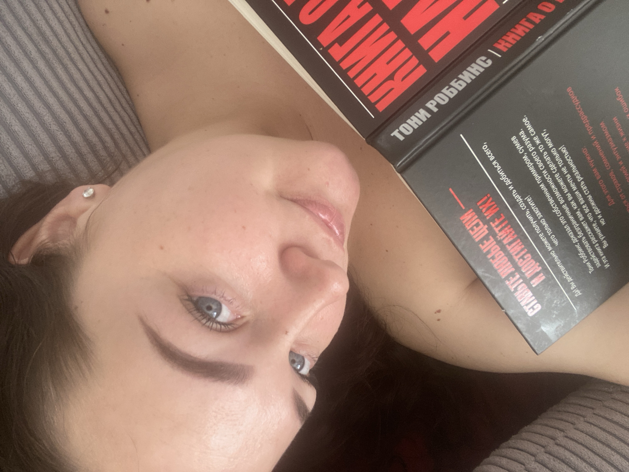 The image shows a woman lying down, holding a book in front of her. The book is "Книга о Тони Роббинсе" (A Book About Tony Robbins) in Russian. The woman is looking at the camera with blue eyes. She has dark hair. An earring is visible in her ear. She is lying on a grey striped cushion. There is no visual information to determine the location in the city of Sochi, Russia.