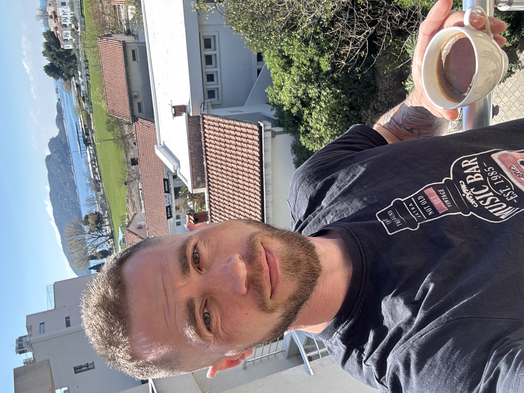 A man with a beard is holding a mug of coffee on a balcony in Budva, Montenegro. The view includes tiled rooftops, green space, a body of water with mountains in the background, and buildings. The man is wearing a black T-shirt with a graphic print. He has a tattoo on his arm. The buildings in the background consist of residential and commercial properties. The photo appears to be taken during the day with good lighting.