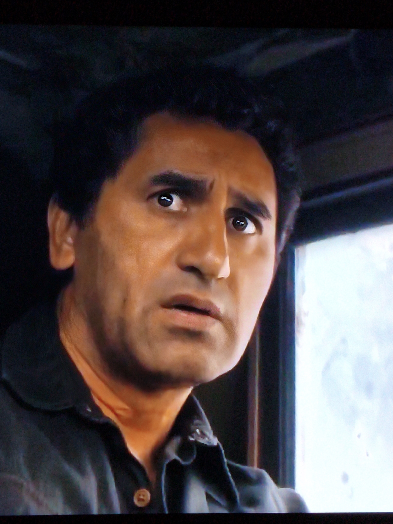 The image is a close-up shot of a man's face. He has dark hair, brown eyes, and a light brown complexion. His mouth is slightly open, and his eyes are looking off to the side with a concerned or surprised expression. He is wearing a dark collared shirt with a single button visible at the collar. The background is dark and out of focus, with a blurred bright area to the right, possibly indicating a window or light source. There is no discernible information about the location in Vladivostok, Russia.