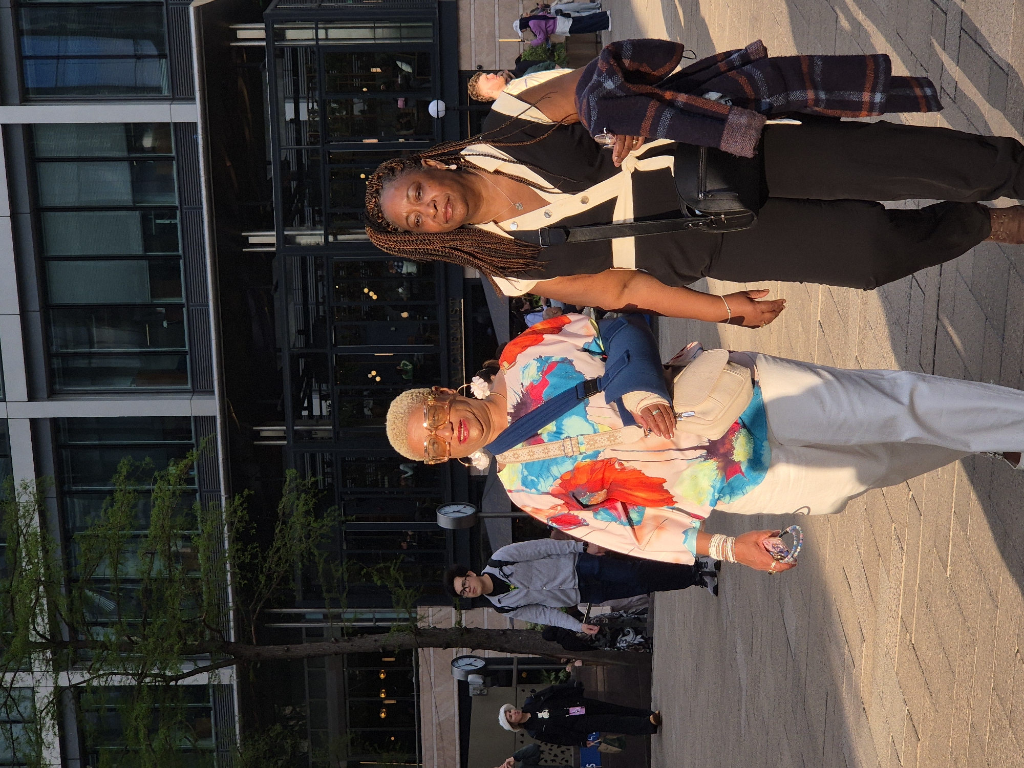 Two women stand on a paved surface in front of a building. The building features dark glass windows and is partially obscured by a tree on the left side.
The woman in the foreground wears a vibrant, patterned top, white pants, sunglasses, and jewelry, and carries a light-colored purse. She holds an item in her hand.
The woman behind her wears a black and white top, black pants, and a patterned jacket draped over her arm. She also carries a black purse.
Other people walk in the background.