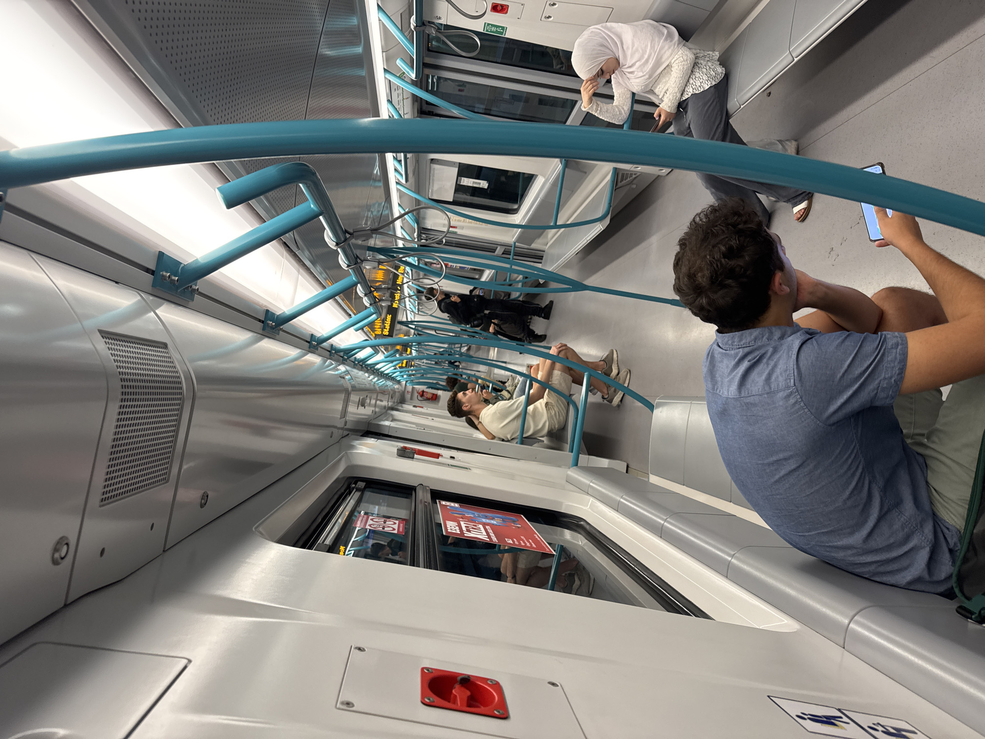 This is a photograph taken inside a subway train in Sofia, Bulgaria. The train appears to be modern and clean, with white and silver interior panels, blue handrails, and gray seating. The image captures a variety of passengers, some seated and others standing. One passenger in the foreground, a young man with curly hair, is seated and looking at his phone. Further down the train, other passengers are visible, including a woman wearing a hijab and a man in a black jacket. The lighting suggests it is daytime. The overall impression is of a typical commute on public transport.