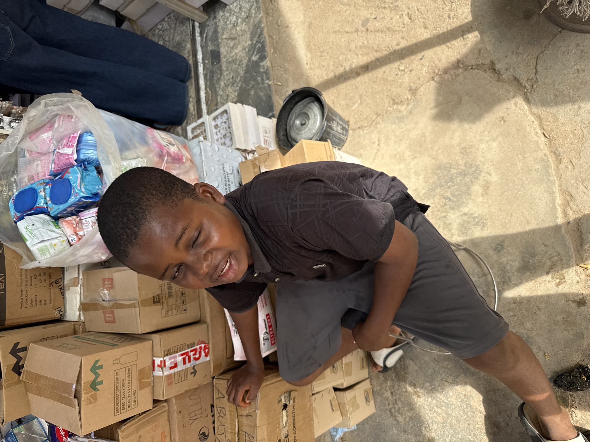 A high-angle, full shot captures a young person, possibly a child, smiling and partially obscured by stacks of cardboard boxes and other items. The person is wearing a dark short-sleeved patterned shirt and gray shorts, with visible footwear on their feet. Their skin tone is dark.
The setting appears to be a cluttered outdoor area, possibly in a commercial or market environment, characterized by a mix of cardboard boxes, plastic packaging, and other containers. Some boxes are labeled with details such as "SKIN PERFECT TONE BODY OIL", "YELLOW", and "ULTRA DRY", indicating a business or trade context. A black bucket and some white boxes are also present.
The scene's location is in the city of Abuja, Nigeria, although this is implied through context as opposed to being directly stated in the image. The ground is a mix of concrete and dirt, with some debris scattered around. Overall, the image conveys a sense of activity and commerce.