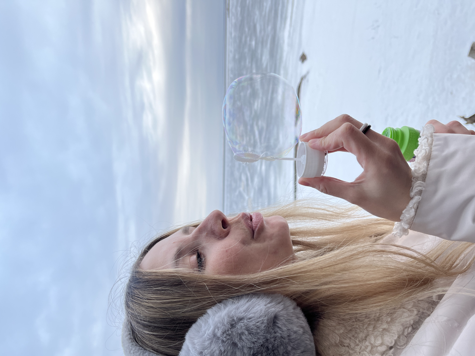 A woman stands outdoors in Chaikovsky, Russia, blowing bubbles. She wears a white jacket and a grey fur hat, and has blonde hair. She holds a green bubble solution bottle and a white wand. A large bubble floats in front of her, partially obscuring the background which includes a body of water and a cloudy sky. She is holding a bubble wand that is black and white.