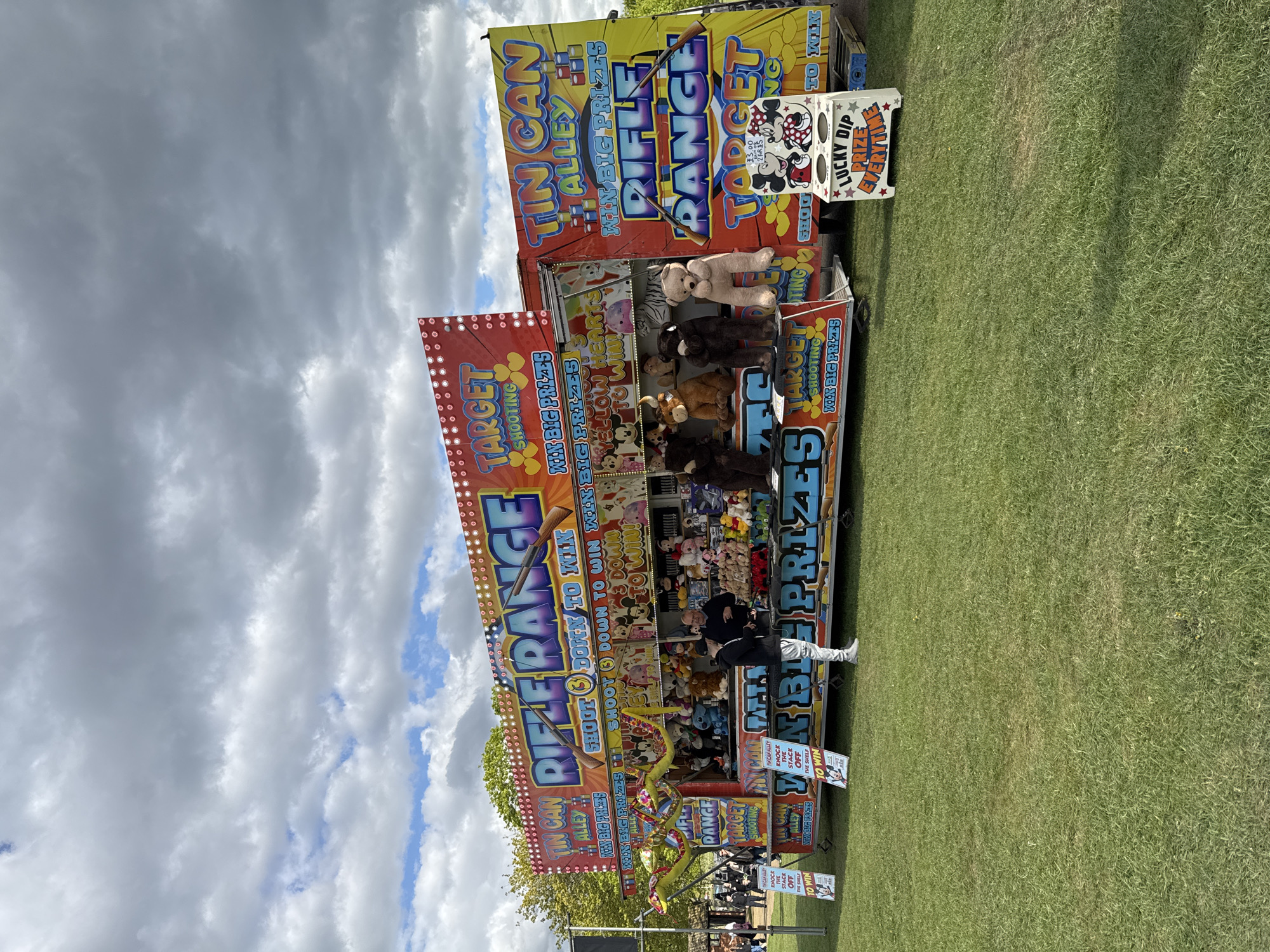 The image captures a mobile carnival game booth, offering a "Rifle Range" and "Tin Can Alley" game, set up on a grassy field under a cloudy sky. The booth is decorated with colorful signage advertising the games and prizes available, with stuffed animals and other prizes displayed. A person stands at the booth, seemingly engaging with the game. The location appears to be an outdoor event or festival. Though the city and country are known to be in South Norfolk, United Kingdom, the picture does not show specific elements that would confirm it.