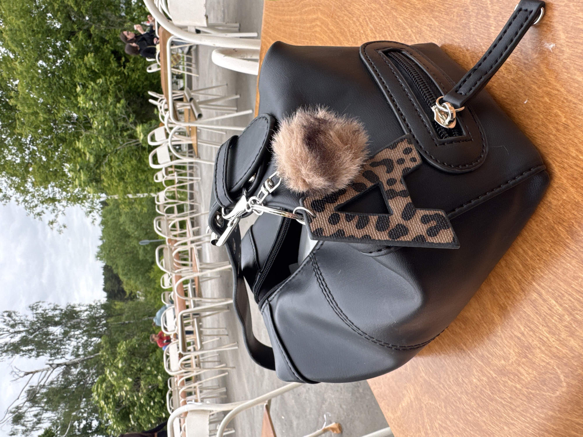 The image primarily features a black, possibly faux-leather, handbag placed on a light brown wooden table. The bag exhibits clean stitching and includes a small zippered pocket on the side, fastened with a silver-toned zipper pull. Attached to the bag are two keychains: a fluffy, brown and black patterned pom-pom, and a flat, leopard-print charm clearly shaped like the capital letter "A".
In the blurred background, an outdoor cafe or restaurant setting is visible. Numerous white metal chairs and matching tables are arranged across a paved area. A few indistinct individuals can be seen seated at some of the tables. Beyond the cafe, a dense collection of green trees and foliage stretches under a cloudy, overcast sky.
The scene takes place during the daytime, characterized by soft, diffused light due to the cloud cover. The wooden table has a visible grain, suggesting natural material. The setup of tables and chairs indicates a public outdoor dining area, possibly within a park or a city square. There are no discernible emotions conveyed. The only visible text is the letter 'A' on the leopard-print keychain. This outdoor cafe setting is located in Moscow, Russia.