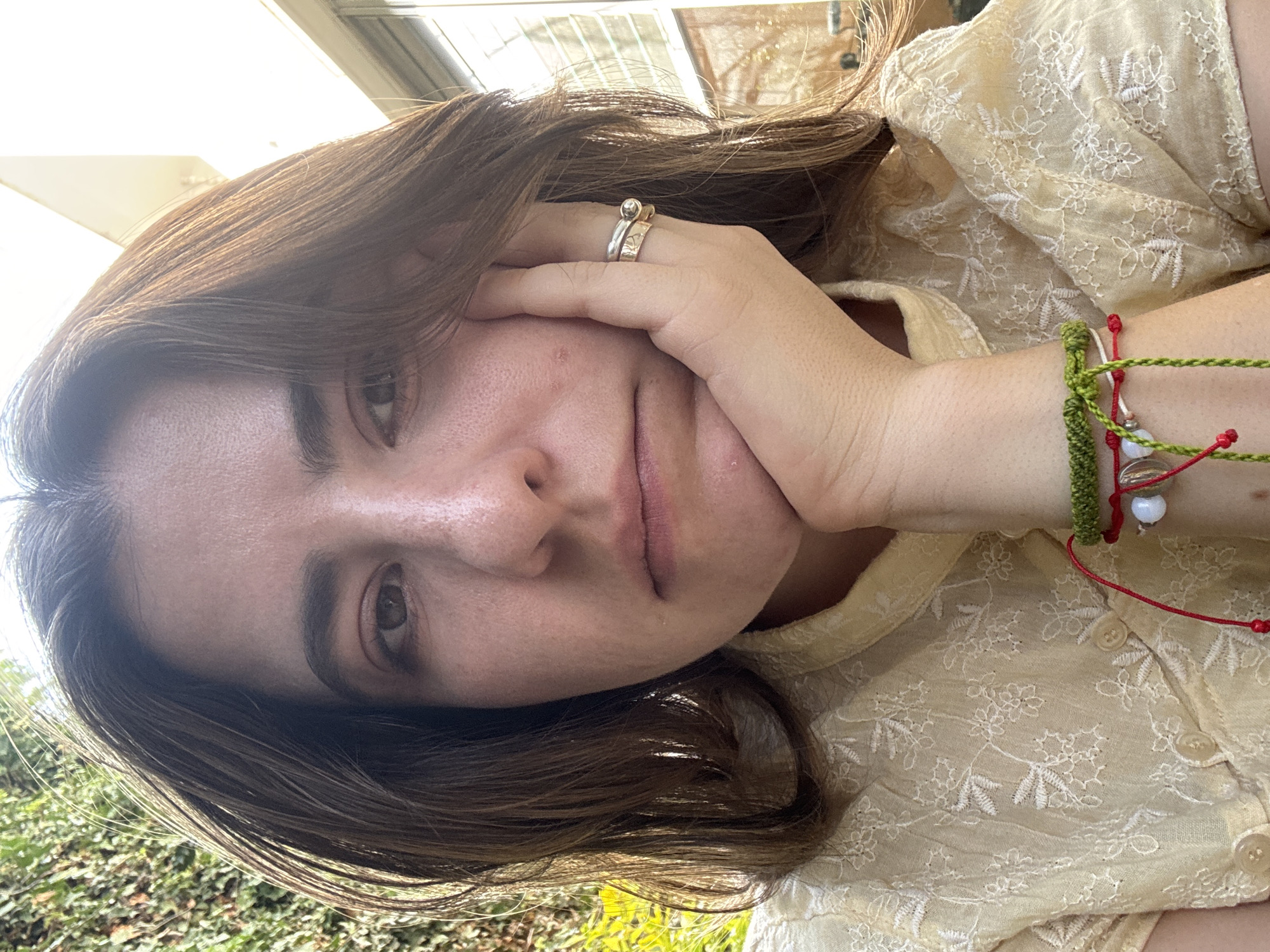 A young woman with light brown hair and fair skin is seen in a close-up, tilted shot. She is wearing a pale yellow, embroidered top and has several bracelets on her right wrist and rings on her fingers. Her right hand is resting against her face. In the background, there is greenery. Given the presence of greenery and architectural styles often found in Puebla, Mexico, it's plausible she's outdoors in a garden or patio setting within the city, but precise location specifics are not identifiable from the provided image.