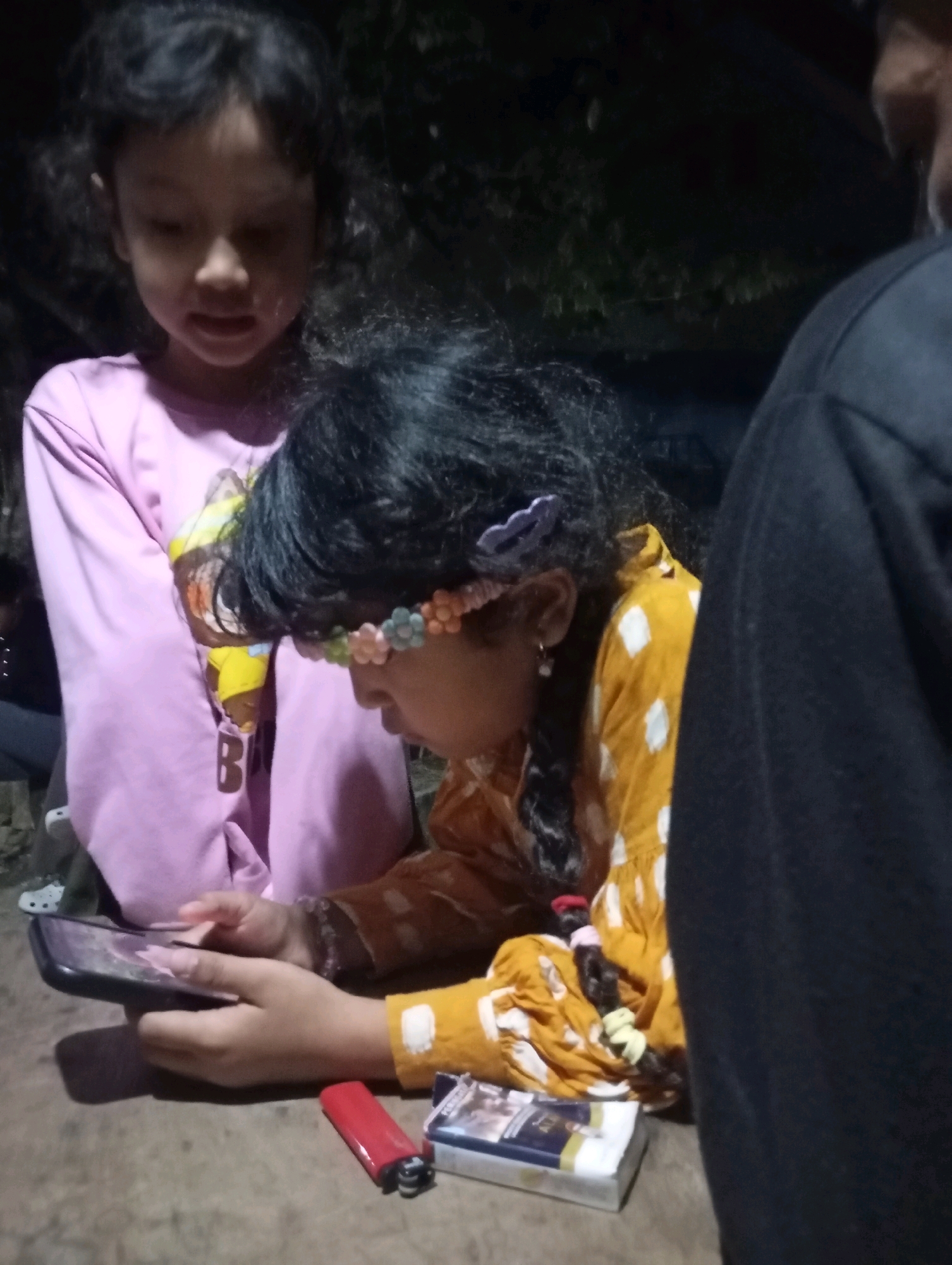 In Ketepung, Indonesia, two young girls are engaged with a smartphone on a wooden surface, likely a table. The girl on the left, wearing a pink long-sleeved shirt with a graphic print, is positioned behind and to the left of the girl on the right. The girl on the right, adorned with a floral headband, wears a yellow and white patterned long-sleeved garment and is actively using the smartphone, holding it with both hands. Adjacent to the smartphone lies a red lighter and a pack of cigarettes. The background is dark and indistinct, suggesting an outdoor or dimly lit environment. A portion of another person's dark clothing is visible on the far right of the frame.