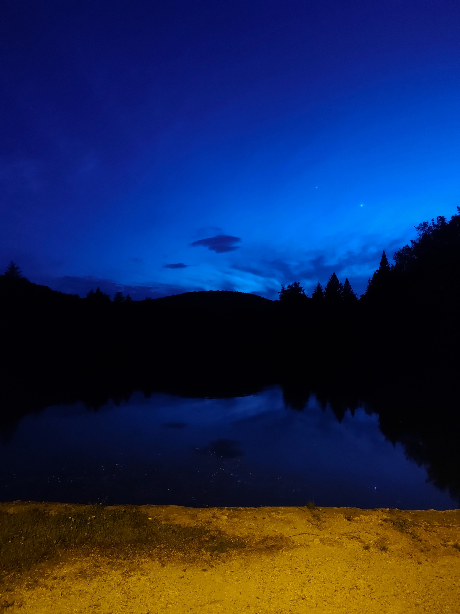 This nighttime image captures a serene natural landscape, likely in Starozagorski bani, Bulgaria. The scene is dominated by a deep blue sky transitioning from a luminous, almost electric blue near the horizon to a darker, intense indigo overhead. Scattered dark clouds are visible, and a few faint stars or planets dot the upper sky.
Below the sky, the silhouettes of distant hills or mountains form a dark, undulating horizon. In front of these, a dense forest of trees appears as an almost entirely black mass, their forms reflected, though somewhat indistinctly, in the calm body of water that occupies the mid-ground. This water, likely a lake or pond, mirrors the dark landforms and the profound blue of the sky above.
The immediate foreground is brightly illuminated by an unseen artificial light source, casting a strong yellow-orange glow on the ground. This area appears to be a dirt path or bank, with sparse patches of green grass and small flora visible under the stark light. The strong contrast between the vibrant, artificially lit foreground and the dark, natural backdrop creates a dramatic and peaceful atmosphere. No people or specific activities are visible, emphasizing the tranquility and stillness of the night. There is no visible text in the image.