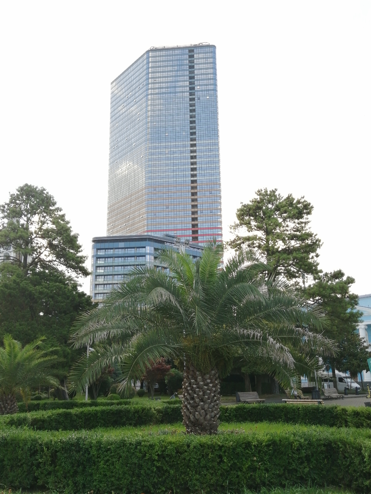 The image shows a modern skyscraper in Batumi, Georgia, featuring a glass facade that reflects the sky. In the foreground, a lush green park with manicured hedges and palm trees creates a vibrant contrast to the urban architecture. Benches are scattered throughout the park, suggesting a public space for relaxation and enjoyment. The background hints at a bright, possibly overcast sky.