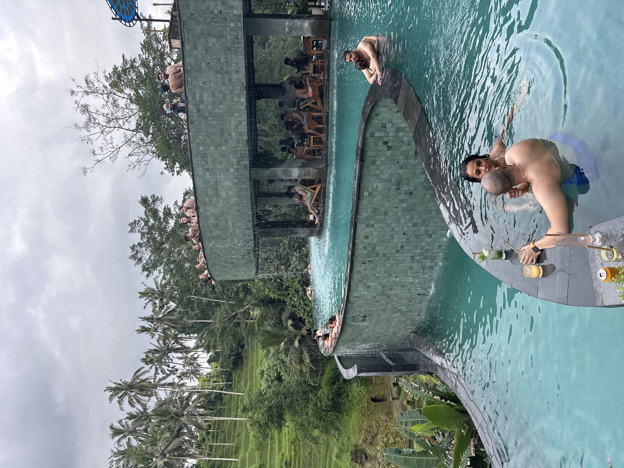 The image depicts a multi-tiered infinity pool at a resort in Tegallalang, Indonesia. Lush, green rice terraces and palm trees fill the background, beneath an overcast sky. People are visible in and around the pools. A man and a woman are partially submerged in the pool in the foreground. Several drinks and cans are set on the pool edge next to them. Other people are seated in chairs and lounging on the upper tiers of the pool complex. A waterfall feature connects the tiers.