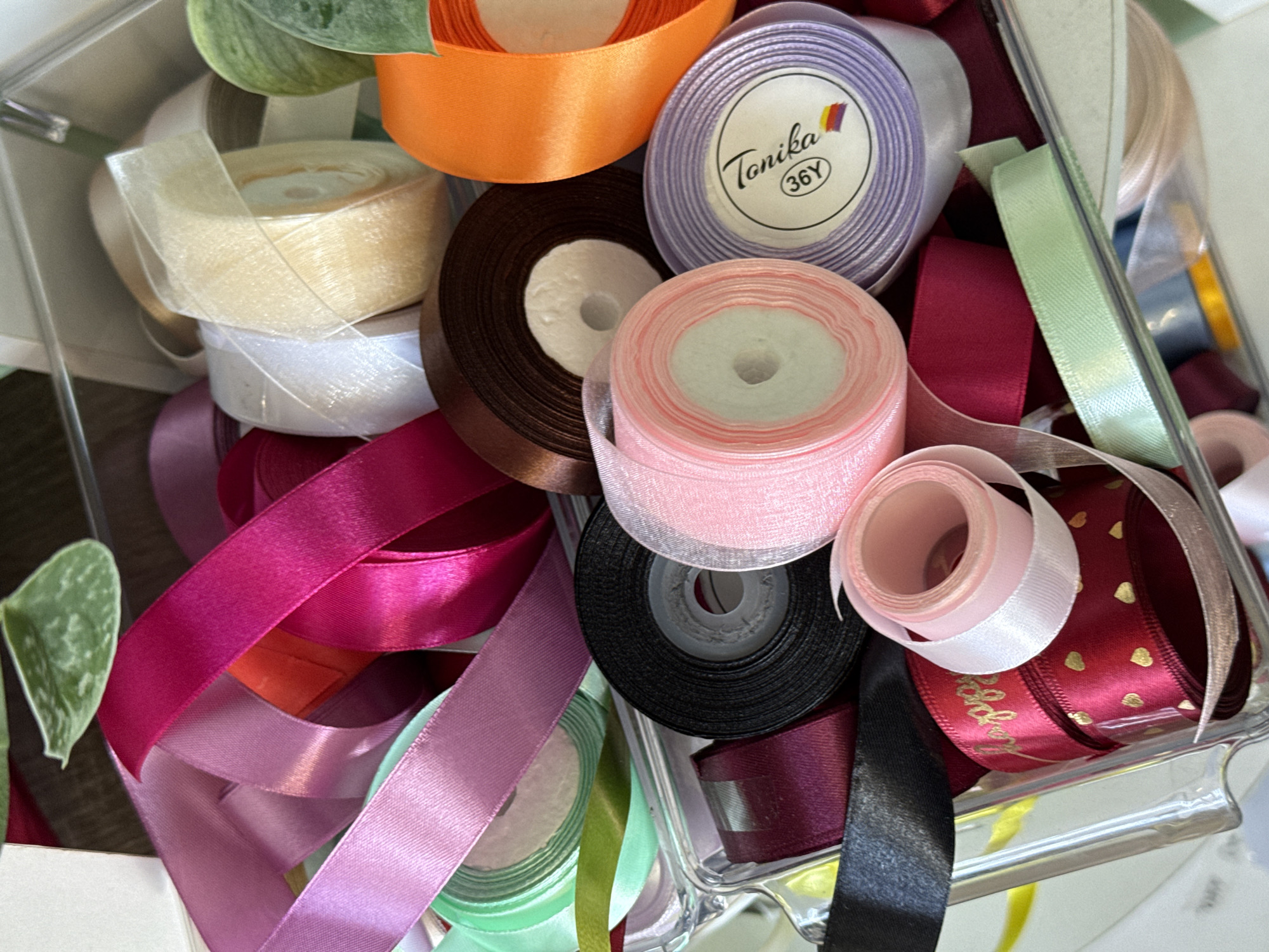 A close-up, overhead shot showcases a clear plastic container filled with spools of colorful ribbon. The ribbons are predominantly in shades of pink, purple, red, green, white, and orange, with a few spools in brown and black. Some ribbons are made of satin, while others appear to be sheer organza. One spool of purple ribbon has a white label with "Tonika" and "36Y" printed on it. The ribbons are arranged in a somewhat jumbled fashion within the container, with some spools peeking out. The background is blurred, but appears to be a wooden surface. There are no people or animals present. The image does not provide any information about a specific city or location.