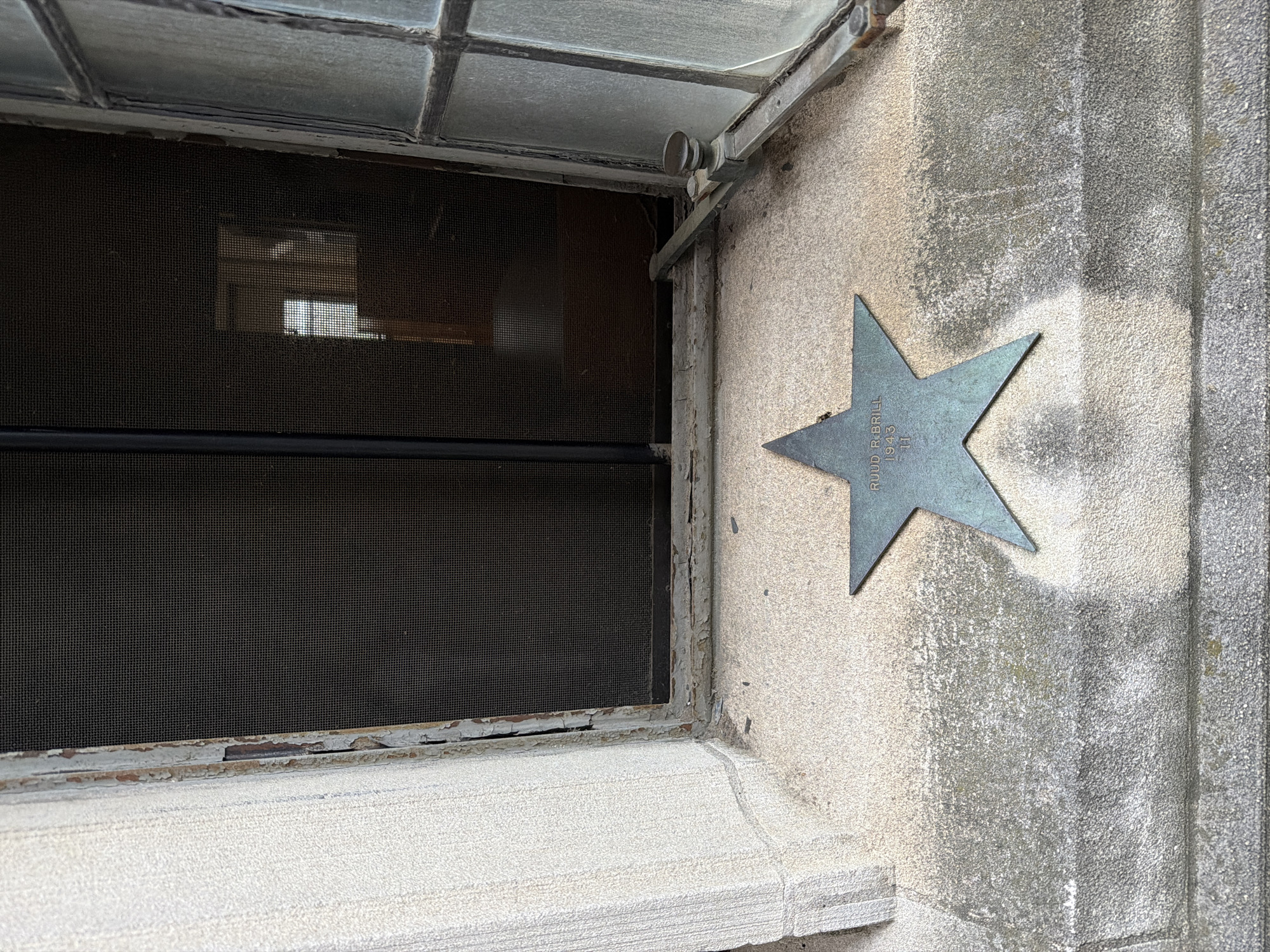 The image shows a bronze star plaque embedded in a stone ledge below a window. The plaque is star-shaped and has text engraved on it, which appears to read "RUUD R. BRILL 1943 11". The window above has a dark mesh screen and a slightly ajar casement window with a metal latch. The stone ledge and the surrounding wall are weathered, with some discolored patches and what appear to be small debris. In the background, through the window, a dimly lit interior with another window can be seen. This plaque is located at Princeton University, specifically on the exterior of the Graduate College.