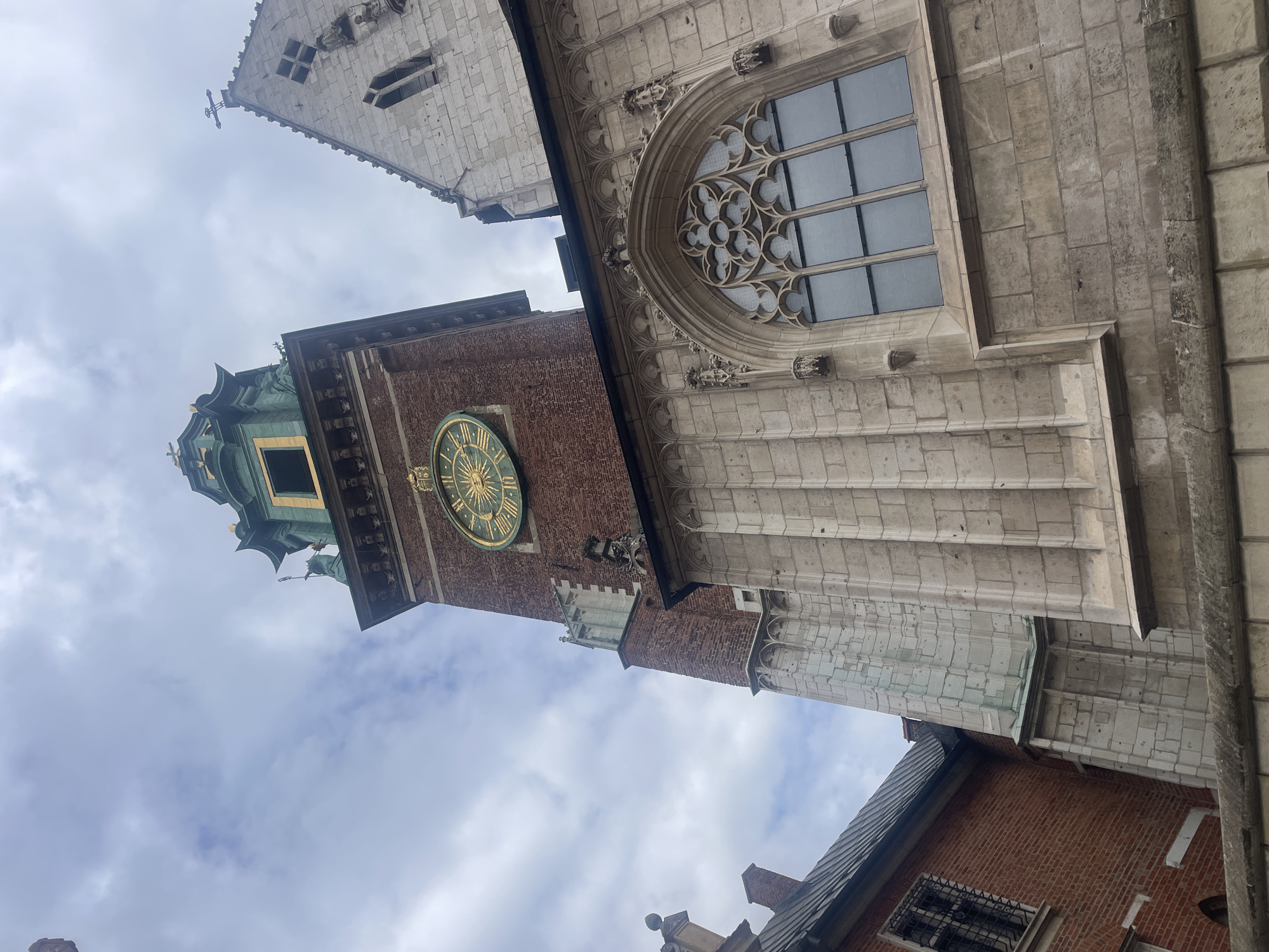 The image captures a low-angle view of the Wawel Cathedral tower in Krakow, Poland. The tower is constructed of red brick and features a large, ornate clock face with Roman numerals and a sunburst design. A green spire crowns the top of the tower. Adjacent to the brick tower is a section of the cathedral made of light-colored stone, showcasing Gothic architectural elements like pointed arches and intricate carvings. A large, multi-paned Gothic window is visible on the stone facade. The sky above is overcast with scattered clouds. The scene is devoid of people.