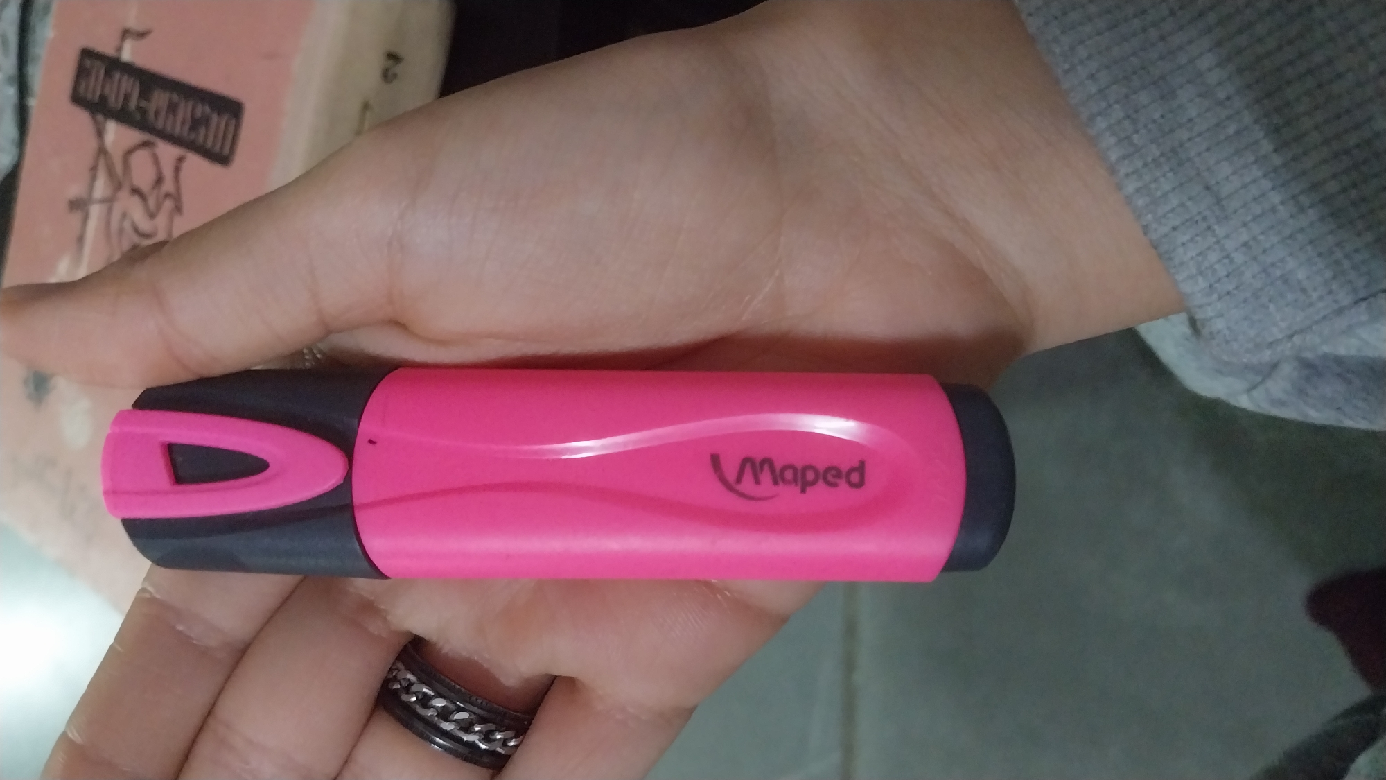 A person's left hand, with light skin tone, holds a pink and black Maped brand highlighter pen in a vertical orientation. A metal ring with a chain-link pattern is present on the ring finger. The person wears a grey long-sleeved garment, partially visible as a cuff in the lower right. In the blurred background, an open or partially open book with a light peach-colored cover displays text in Armenian script, including "ՄԱՐԴ-ԱՄԵՆ" and the numeral "2". The scene takes place indoors, inferred to be in Yerevan, Armenia, due to the visible Armenian text on the book.