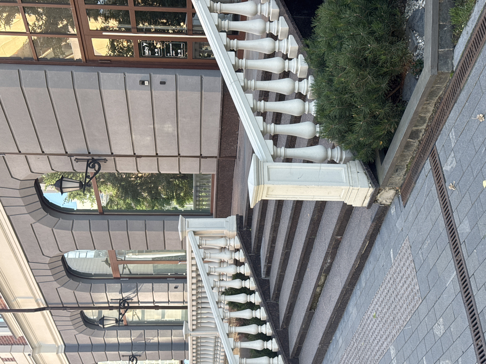 The image depicts an outdoor scene featuring an ornate white balustrade alongside a set of stairs. The balustrade has a classic design with repeated decorative balusters. To the left of the balustrade is a building with a gray, paneled facade and large windows. Some windows have reflections of trees and the sky. A black wrought-iron lamp with a lantern is attached to the building's facade. To the right of the stairs is a dense green shrub. The stairs are made of dark stone with lighter gray treads. The ground next to the stairs is paved with gray square tiles, and a metal grate runs along the edge.
In the background, through a window of the building, a sign is partially visible that reads "saby" followed by "9-18" twice, suggesting business hours. The lighting suggests it is daytime, possibly late afternoon, with the sun casting shadows and creating reflections on the windows. The overall impression is one of an exterior entrance to a building, possibly a commercial establishment or a well-maintained public space, with a focus on architectural details. The setting appears to be Yekaterinburg, Russia. There are no people visible in the image, and the scene is static.
