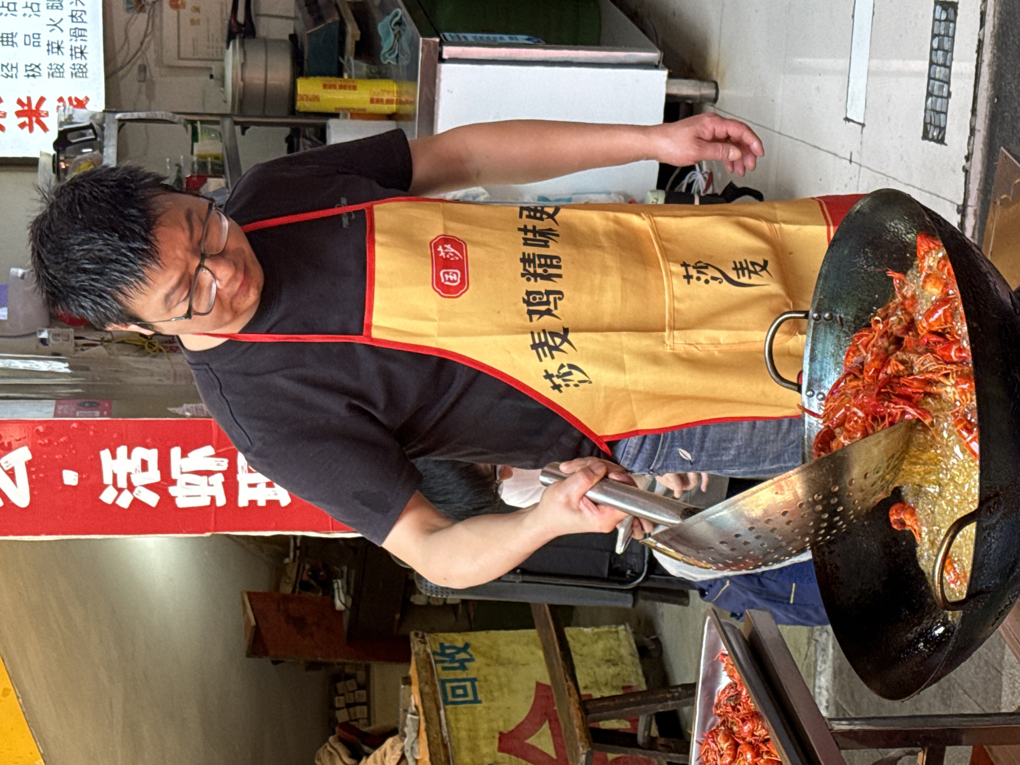 A vendor wearing a yellow apron with red trim and the words "莎麦鸡精味" (Sha Mai Chicken Essence Flavor) fries crawfish in a large wok on a street in Beibei, China. He uses a slotted spoon to remove the crawfish and place them on a metal tray. A red sign in Chinese characters is displayed nearby, and there is a green and yellow wooden sign that says “回收” (Recycle) on the bottom left of the photo.