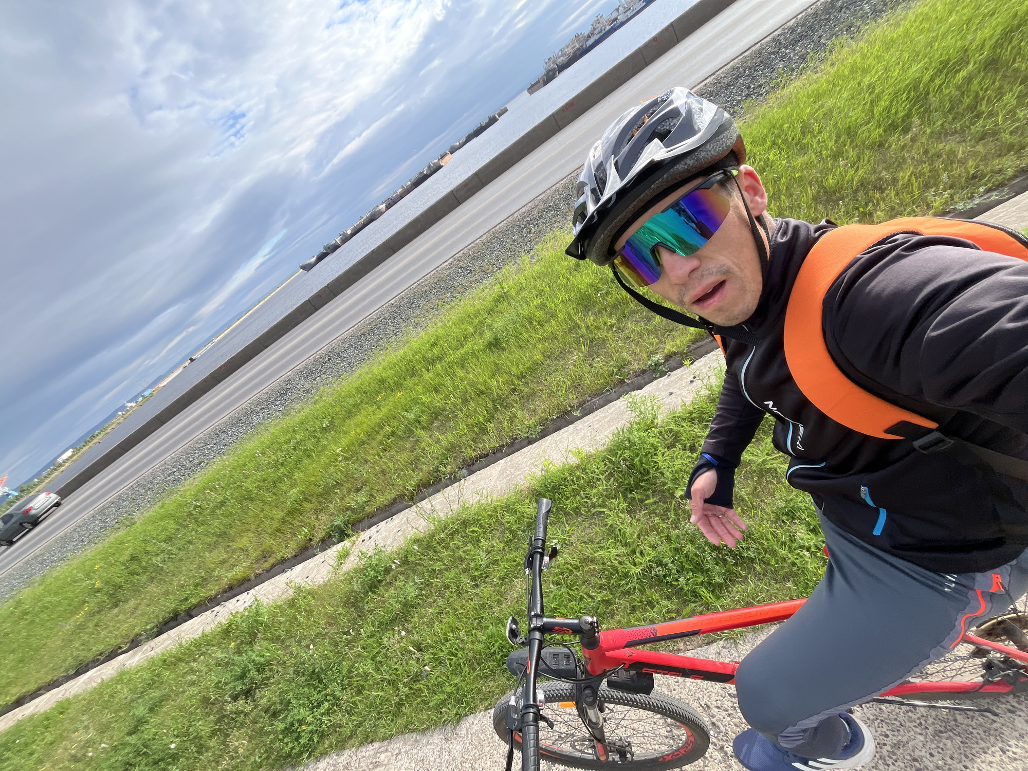 A man wearing a helmet, sunglasses, and athletic clothing is taking a selfie while sitting on a red bicycle. He is positioned on a paved pathway next to a grassy area. In the background, a body of water, possibly the Kama River, is visible with several ships docked. A road with a car driving on it runs parallel to the water. The sky is partly cloudy. The scene appears to be located in Chaikovsky, Russia, along the embankment.