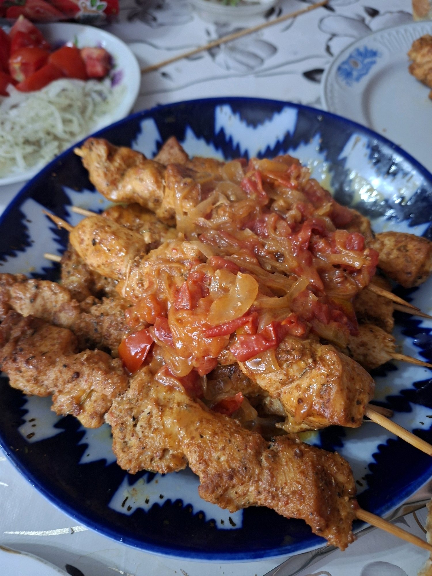 In the city of Manas, Kyrgyzstan, a close-up, eye-level shot captures a platter of chicken skewers topped with a sauce of tomatoes and onions. The skewers, arranged diagonally across the frame, are served on a dark blue plate with a distinctive white zigzag pattern. To the left of the platter, a white plate holds a side dish of sliced tomatoes and thinly shredded onions. The scene is set on a tablecloth with a subtle, abstract design.