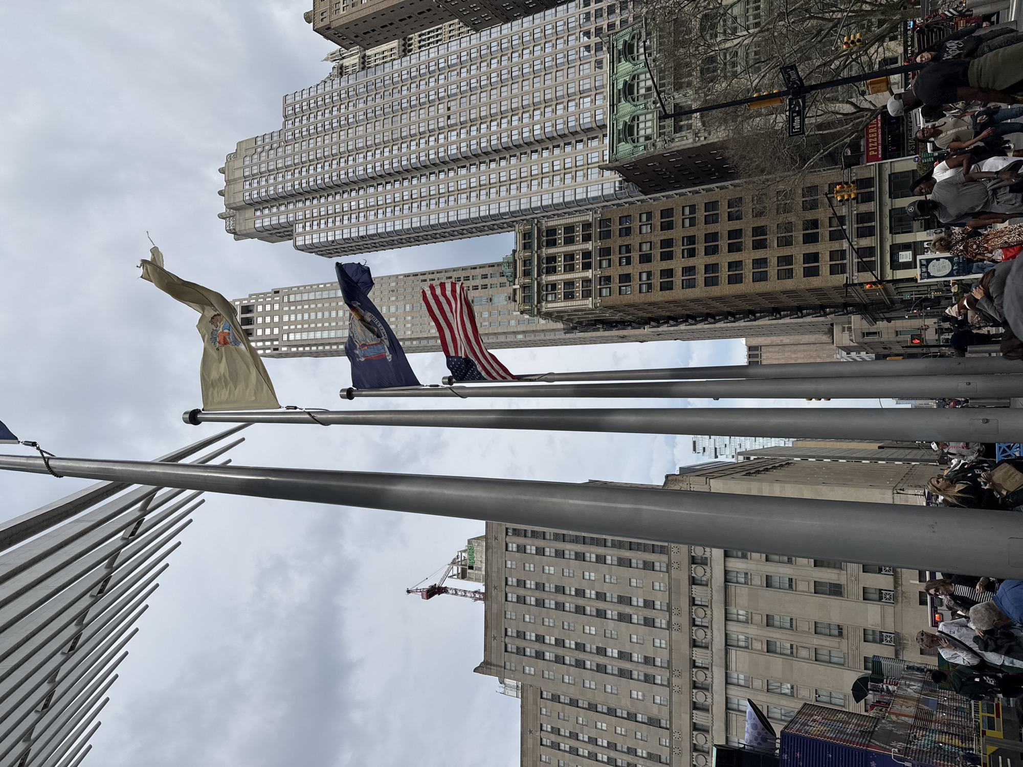 The image shows a street scene in New York City, United States of America, looking upwards towards buildings. Several flag poles are visible, flying the New York City flag, the New York State flag, and the American flag. Tall buildings are present, and people are visible on the street. Street signs labeled "Fulton St." are visible.