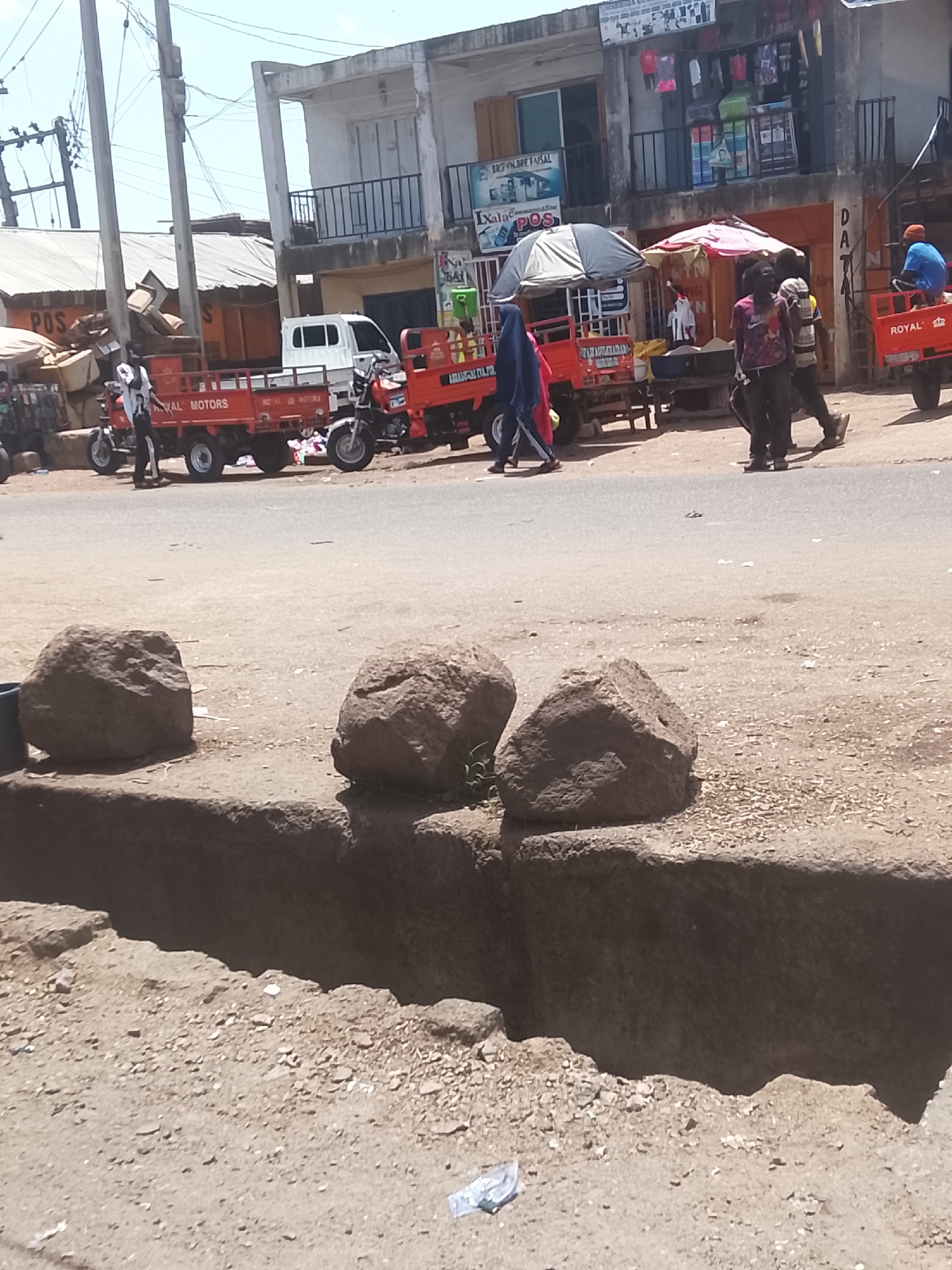 The scene is a street in Jos, Nigeria. The foreground shows a shallow trench with three large rocks resting on the edge. Several red three-wheeled vehicles, some labeled "ROYAL MOTORS" and "ROYAL", are parked on the side of the road. People are walking near the vehicles. Buildings with storefronts line the background. One storefront shows a sign with "BROWN.BRE FAISAL" and "Ixala POS". Another has "AMANTAM DATO" written on it. There's a white truck parked further back. One person is seated on the back of the right most red vehicle.