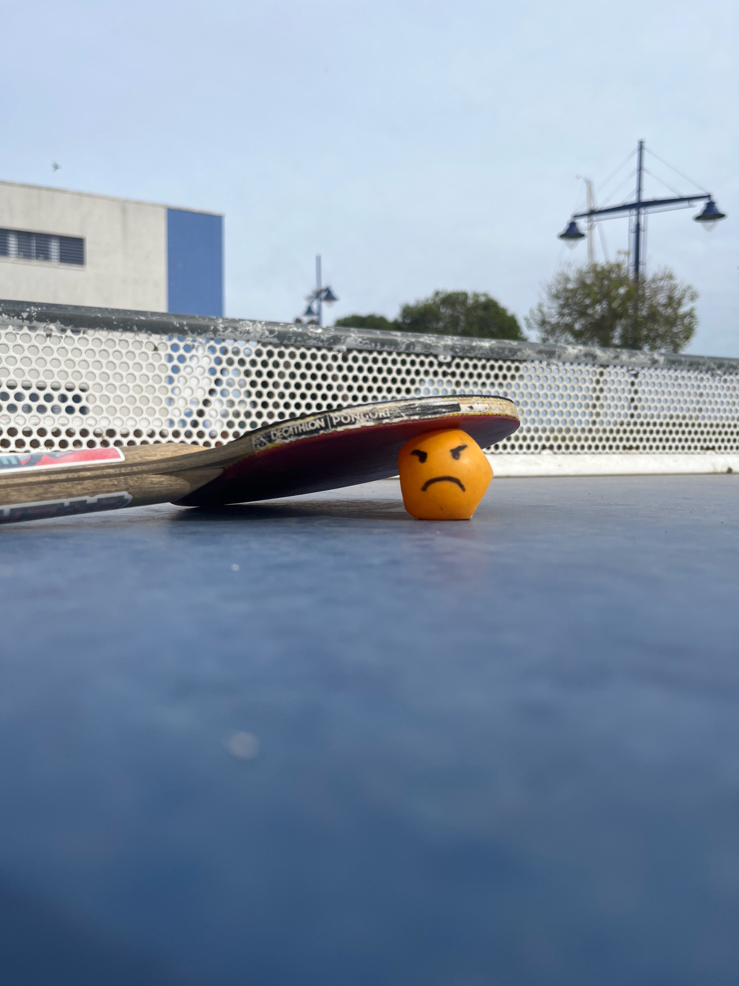 The image shows a close-up, eye-level shot of a blue surface, likely a table tennis table, with a table tennis racket lying on it. The racket's red side is facing up, and it is angled downwards, with its edge resting on an orange table tennis ball. The ball has a simple, hand-drawn angry face on it, with two black dots for eyes and a curved line for a mouth. In the background, a white and blue building with a horizontal window is visible under a cloudy sky. To the right, a street lamp with two lights stands above some trees. The scene appears to be outdoors, potentially in a park or recreational area in Tarragona, Spain, given the architectural style of the building and the presence of outdoor recreational equipment. The overall lighting suggests an overcast day. There are no people visible in the image.