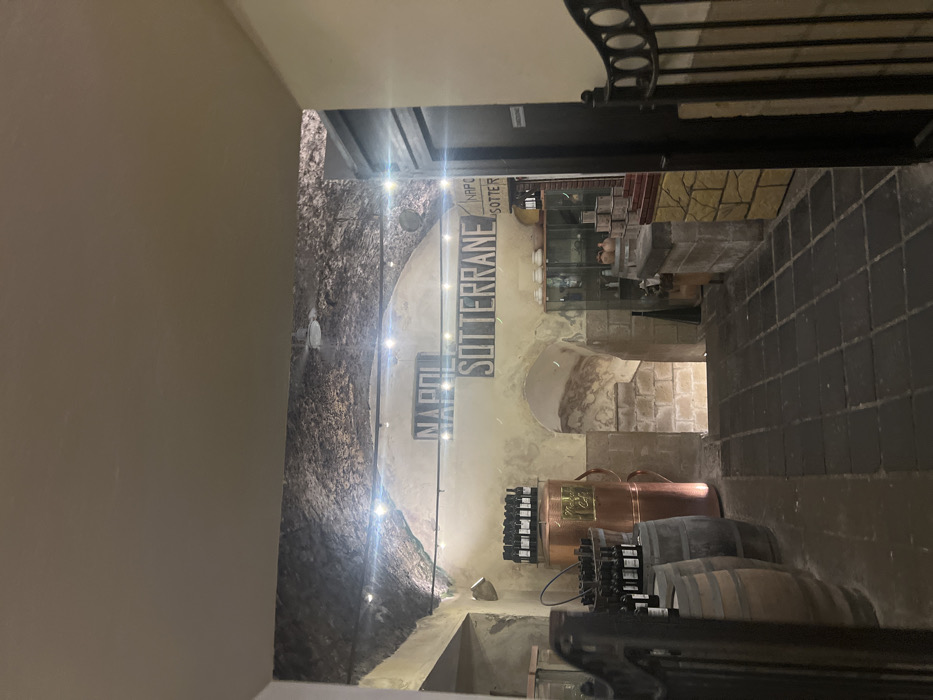 The image shows the interior of a wine cellar, located in Naples, Italy at latitude 40.851 and longitude 14.257. The cellar has a low, vaulted ceiling made of exposed stone. The walls are also made of stone and are painted a pale yellow. The cellar is lit by several overhead lights. On the left side of the image, there are two wooden barrels filled with bottles of wine. Behind the barrels, there is a tall copper vat also containing bottles of wine. On the right side of the image, there is a small, stone counter with a display of wine bottles and other items. Above the counter, there is a sign that reads "Napoli Sotterrane". There is also a black metal gate that leads to an unknown space. The floor is made of tile and is covered in dirt. The cellar is relatively empty, except for the wine barrels, bottles, and the counter. There is no one in the cellar.