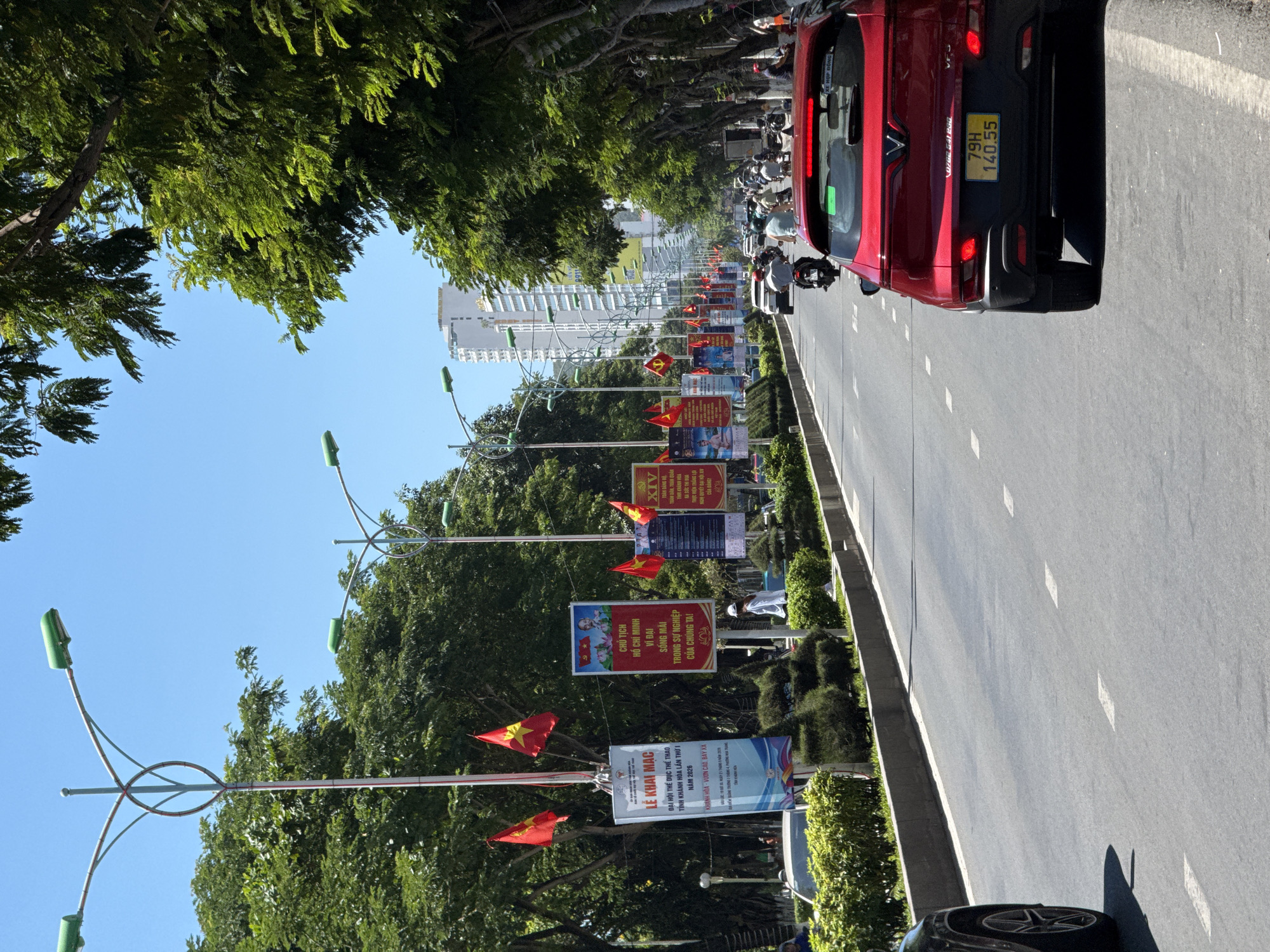 The image captures a bustling street scene in Nha Trang, Vietnam, on a bright, sunny day. The street is lined with lush green trees, their foliage casting shadows on the asphalt. Modern streetlights with green lamps arch over the roadway, adorned with decorative elements. Several Vietnamese flags, red with a yellow star, are prominently displayed on poles along the street, indicating a patriotic or celebratory occasion.
Banners and posters are visible on poles, some displaying text and images. One banner, in Vietnamese, announces "LỄ KHAI MẠC" (Opening Ceremony) and mentions "ĐẠI HỘI THỂ DỤC THỂ THAO TỈNH KHÁNH HÒA LẦN THỨ I NĂM 2026" (Khanh Hoa Provincial Sports Festival 1st Edition Year 2026). Another poster features portraits of Ho Chi Minh and text related to his ideology.
Traffic is moderate, with a mix of vehicles including cars and numerous motorbikes, which are a common mode of transport in Vietnam. A red VinFast car is the most prominent vehicle in the foreground, its license plate reading "79H 140.55". People are visible in the background, some on motorbikes, others walking. A person in a white uniform is standing near a banner.
In the distance, modern high-rise buildings form part of the urban landscape, contrasting with the natural greenery. The overall impression is of a vibrant, active city on a warm day, with elements of local culture and ongoing events evident. The clean asphalt road, marked with white lines, suggests a well-maintained urban environment.