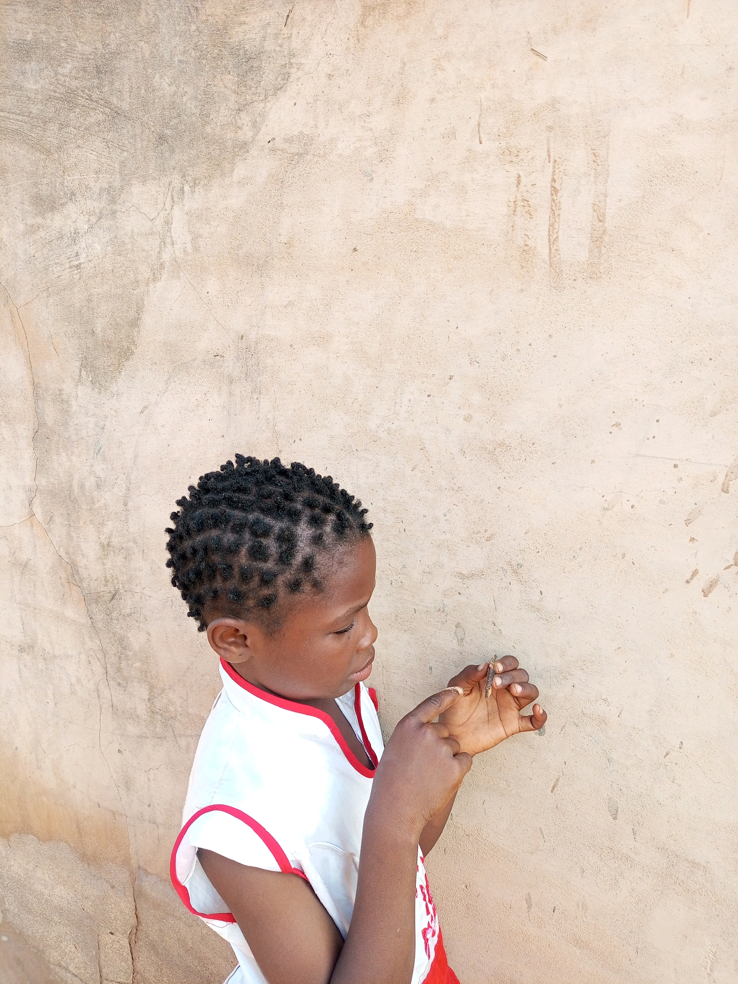 A young child, likely female, stands in profile against a textured, light-brown wall. The child has dark skin and short, braided hair. She wears a white sleeveless shirt with red trim around the collar and armholes. Her right hand is raised, and she appears to be examining something small held between her thumb and forefinger, possibly a twig or insect. Her gaze is directed downwards at her hand, and her left hand is also visible, held near her chest. The wall behind her shows signs of age and weathering, with cracks and uneven patches of color. The scene suggests an outdoor environment, potentially in a rural or less developed area. The location is identified as Tempure, Nigeria.