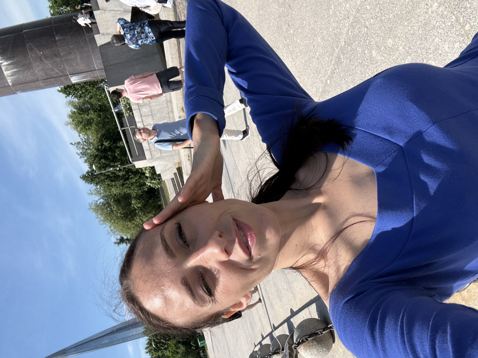 A woman is taking a selfie outdoors in Saint Petersburg, Russia, on a sunny day. She is wearing a blue long-sleeved top and has her right hand on her forehead, her left arm is extended out to the side. In the background, a tall, modern glass skyscraper is visible on the left side of the frame. To the right, there is a large, dark, cylindrical monument or structure. Several people are standing in the middle ground and background, some appearing to be looking at the monument or skyscraper. The ground is paved with light-colored stone or concrete. In the foreground on the left, there is a chain with several large, rounded, gray objects, possibly bollards or decorative elements. Trees and a blue sky with some clouds are also visible.