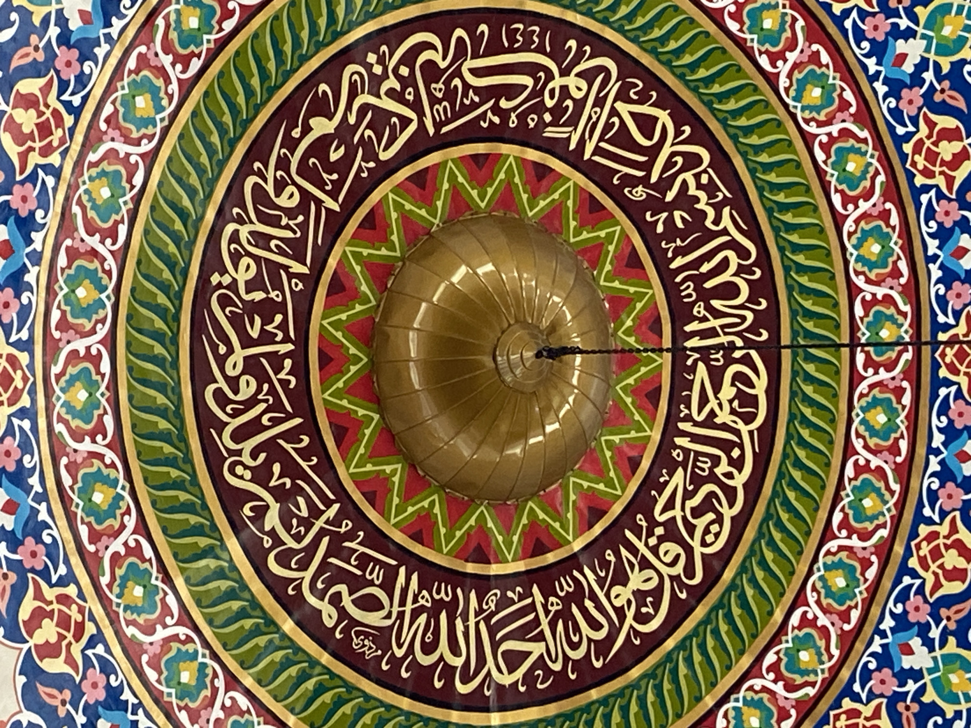 The image shows a close-up, overhead view of a decorated dome's interior, likely from a mosque. The dome itself is a golden, ribbed structure with a hanging cord. Surrounding the dome is a circular motif featuring intricate geometric patterns and Arabic calligraphy in gold lettering against a deep burgundy background. This inner circle is bordered by rings of green, red, and blue, adorned with floral and geometric designs. The style of decoration, particularly the calligraphy and arabesque patterns, is characteristic of Islamic art.
This specific dome, with its prominent Arabic calligraphy and vibrant decorative elements, is a notable feature of the **Masjid Agung Islamic Center Lhokseumawe** in Lhokseumawe, Aceh, Indonesia. The mosque is a significant religious and architectural landmark in the city.