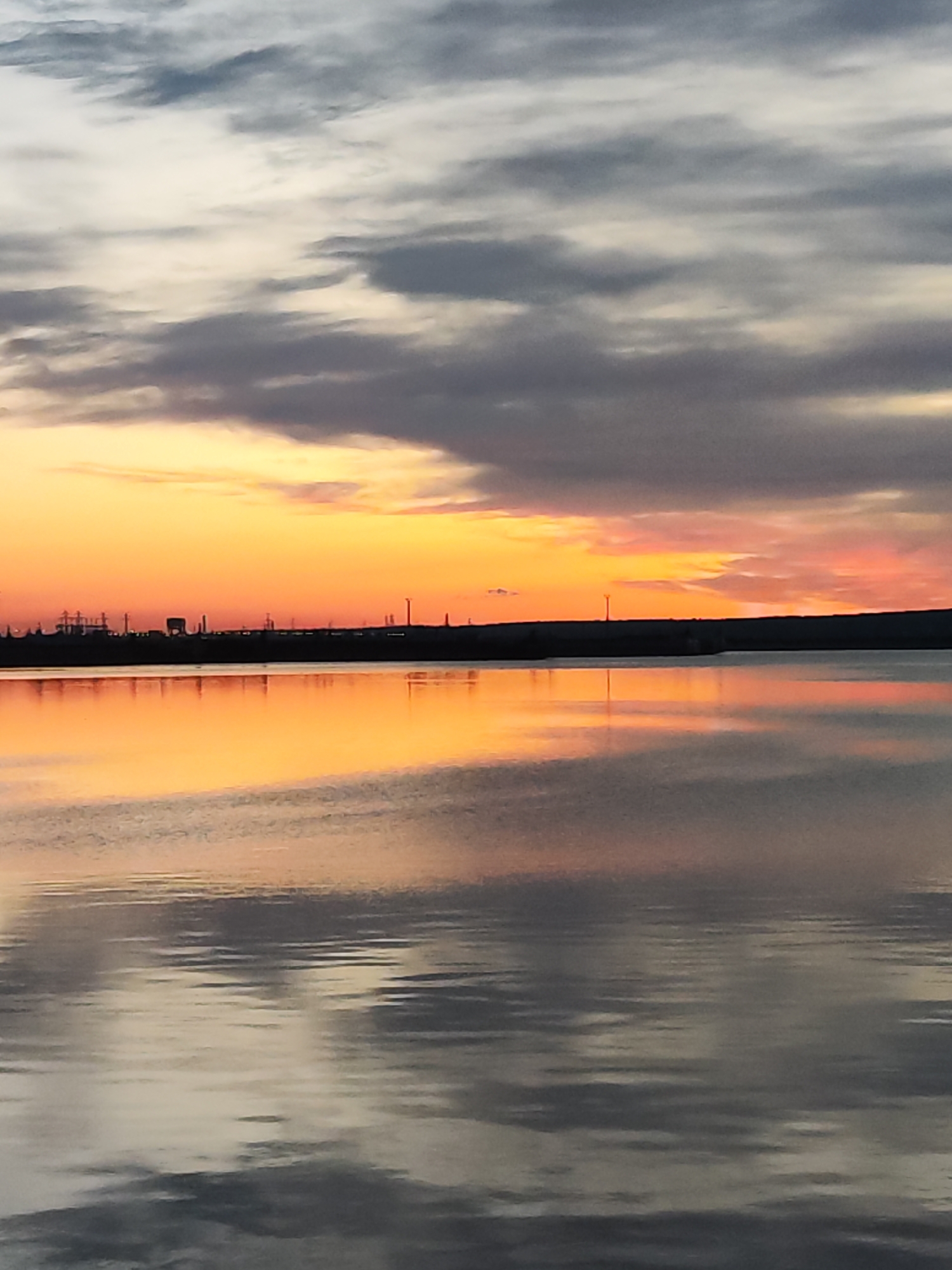 The image captures a sunset over the Kama River in Chaikovsky, Russia. The sky is filled with a mix of orange, pink, and grey clouds. Reflected in the calm water of the river are the colors of the sunset. The silhouette of industrial buildings, including power lines and structures, is visible on the horizon. The light is dim and it appears to be just after sunset. The water has ripples across it.