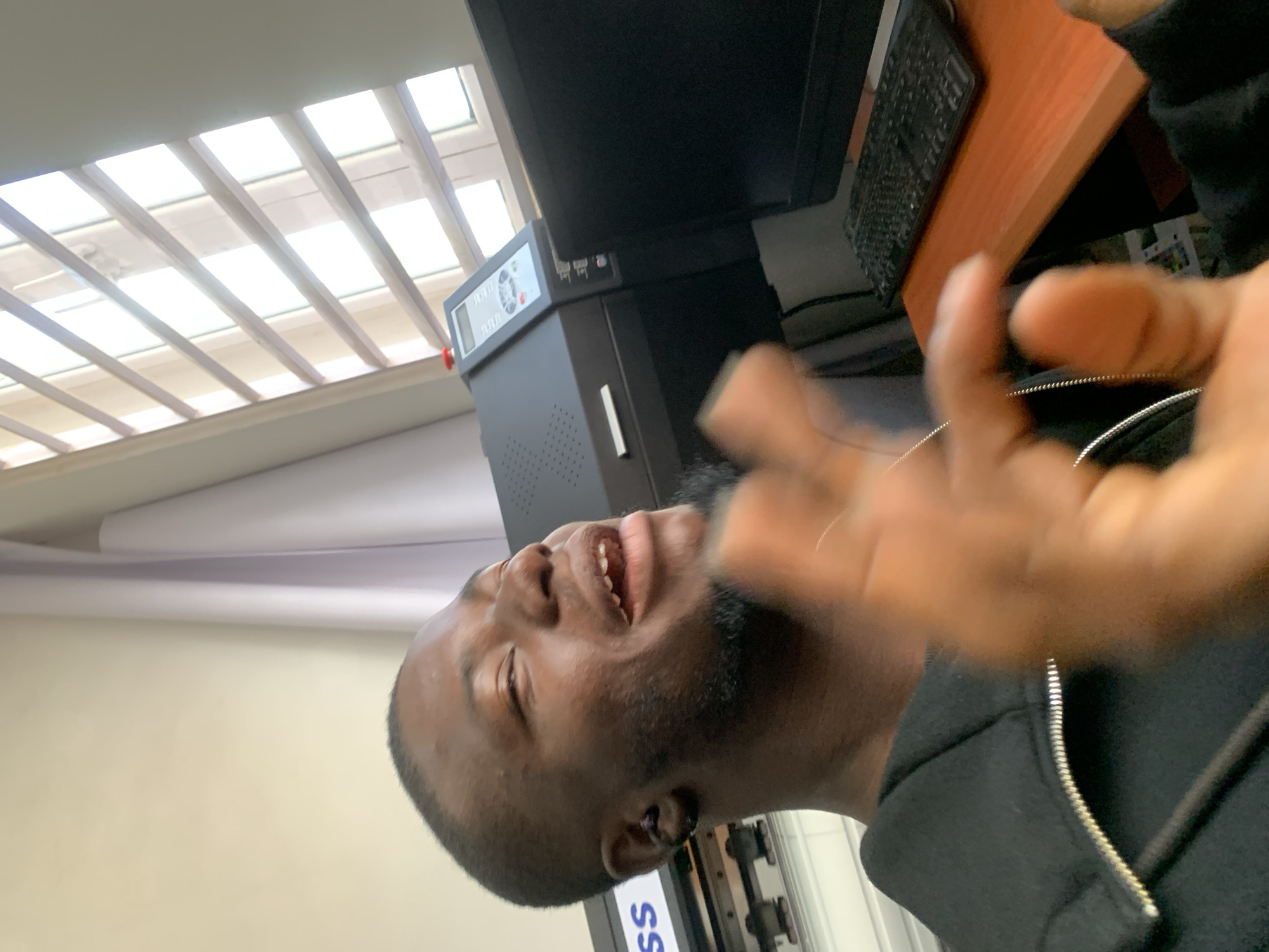 A medium shot captures a young Black man in an office setting, his head and shoulders filling the frame as he gestures with his right hand. He is wearing a black zip-up hoodie and has short, dark hair, a beard, and an earring in his left ear. His eyes are closed, and his mouth is open as if he is speaking or singing. In the background, a black machine with a digital display sits on a desk next to a black computer monitor. The desk has a wooden surface, and a black keyboard is visible. Horizontal blinds are on a window behind the desk, allowing sunlight to filter through. The overall impression is of a candid moment in a work environment.