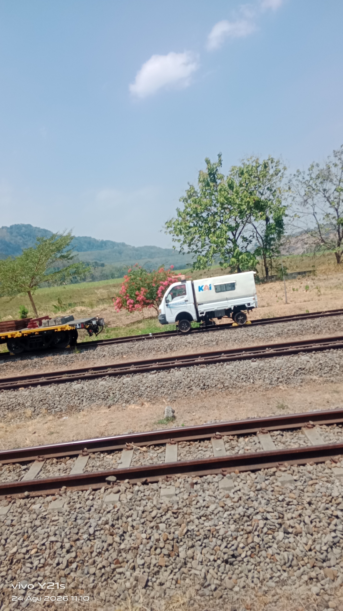 An eye-level, medium shot captures a white, four-wheeled truck with a white cargo box driving on train tracks. The truck has the logo "KAI" in blue and white on its side. In the background, there is a flatbed train car carrying some rectangular objects. A small tree with pink flowers is to the left of the truck. The scene is set in a rural area with dry grass, trees, and rolling hills under a clear blue sky with a few white clouds. The image is a photograph taken on a mobile phone, as indicated by the watermark "vivo Y21s 24 Agu 2026 11.10" in the lower left corner. The location is Prupuk Utara, Indonesia.