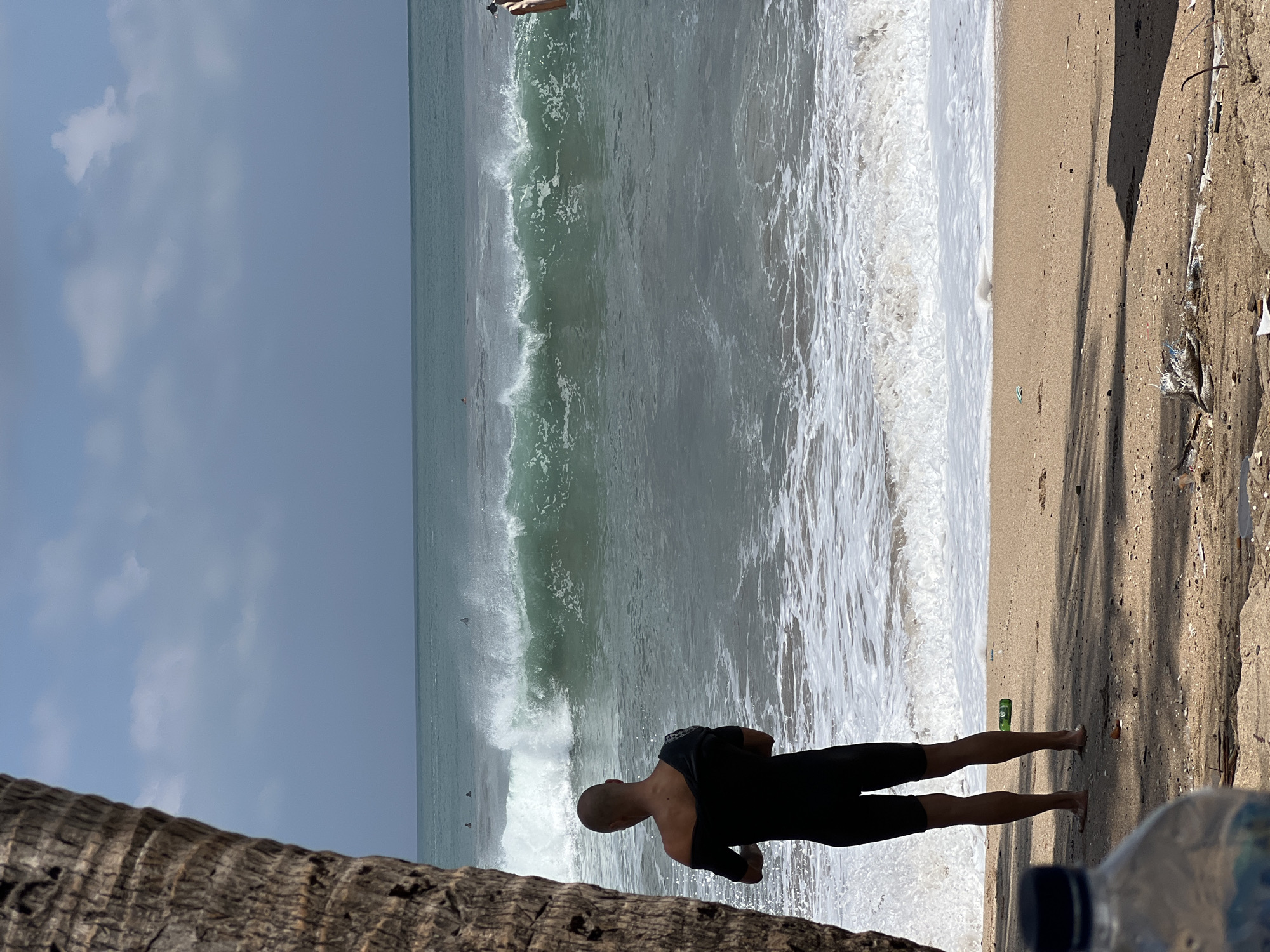 A person is standing on a sandy beach in Kuta, Indonesia, facing the ocean and a large, green wave. The person is wearing a black wetsuit and is in the process of taking it off, with one shoulder exposed. In the background, several surfers are visible in the water, riding or waiting for waves. The sky is partly cloudy, and the sun is casting shadows on the beach, indicating it is daytime. There is a large palm tree trunk on the left side of the frame. A plastic water bottle is visible in the foreground, and some debris is scattered on the sand.