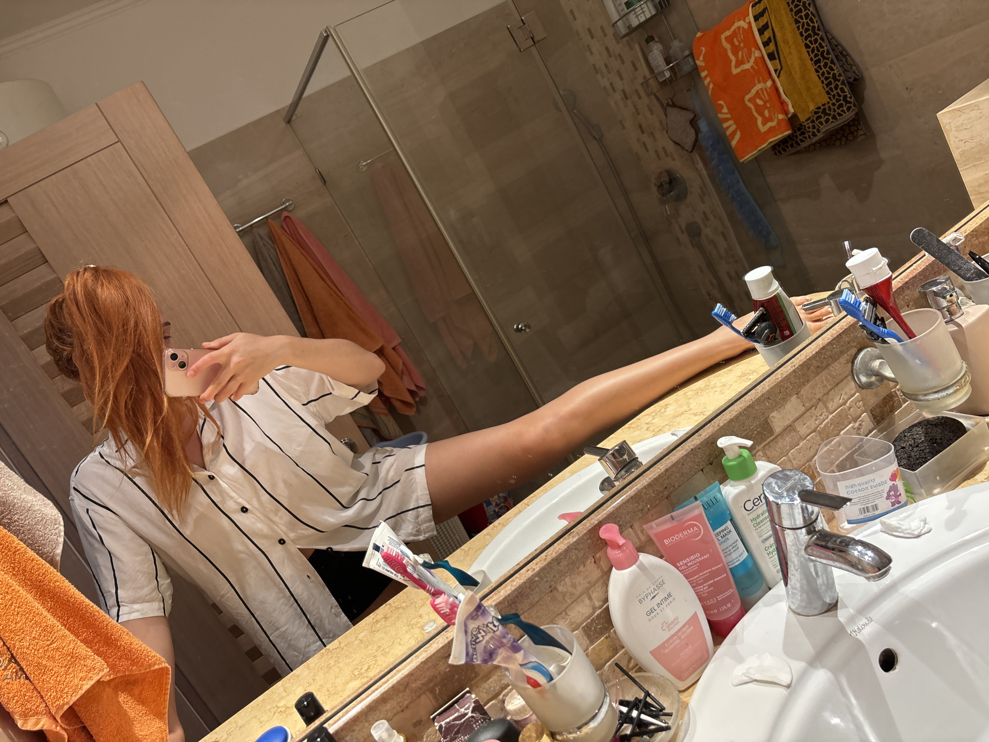 The image depicts a full-length mirror selfie taken in a bathroom. The scene appears to be in Budva, Montenegro, based on contextual information not directly visible in the image. A person with long, auburn hair is holding a pink smartphone and taking a picture of themselves. They are wearing a white shirt with black vertical stripes and dark shorts. The bathroom counter is cluttered with various toiletries, including toothbrushes, toothpaste, lotions, and makeup brushes. A sink is visible in the foreground. In the background, a shower stall with a glass door is present, and several towels are hanging on a rack. The overall impression is of a typical domestic bathroom setting.
