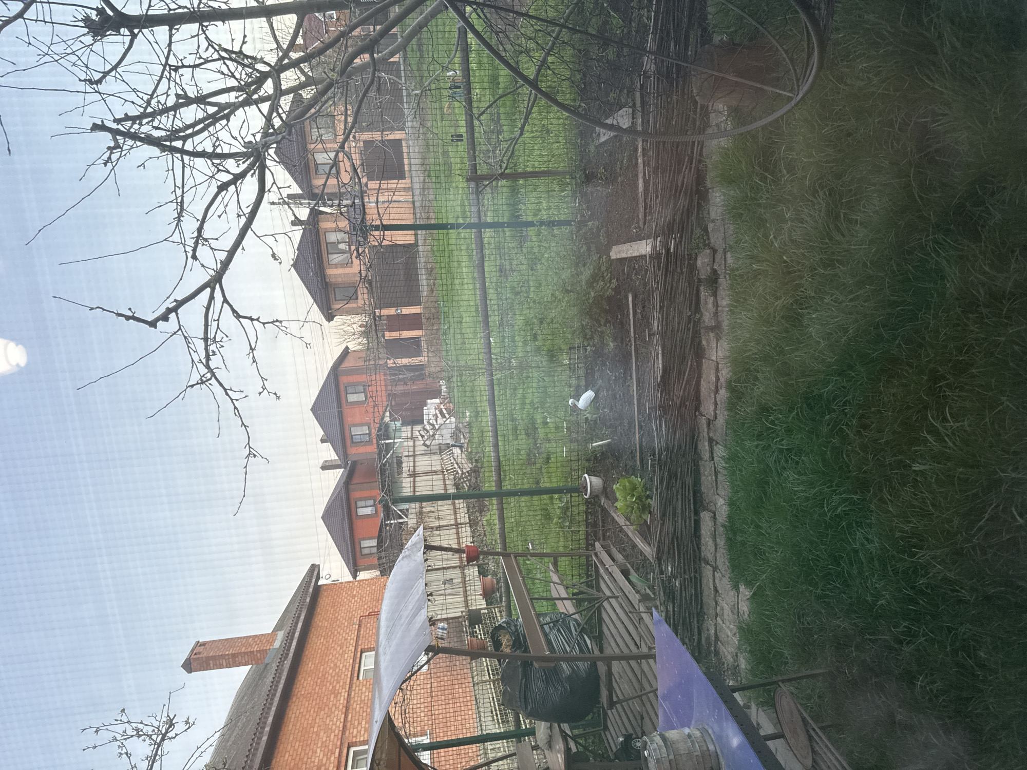 The image shows a view from a window overlooking a residential area. In the foreground, there are bare tree branches on the left and part of a metal structure with blue and clear plastic sheets attached. A black garbage bag hangs on the metal structure. Beyond the structure, there is a fenced garden area with green grass. Further back, several residential houses with red-brick exteriors and brown roofs are visible. The scene appears to be in a suburban or semi-rural area, and the sky is overcast. It is not possible to determine if this scene is in the city of Rostov-on-Don, Russia, with the given information.