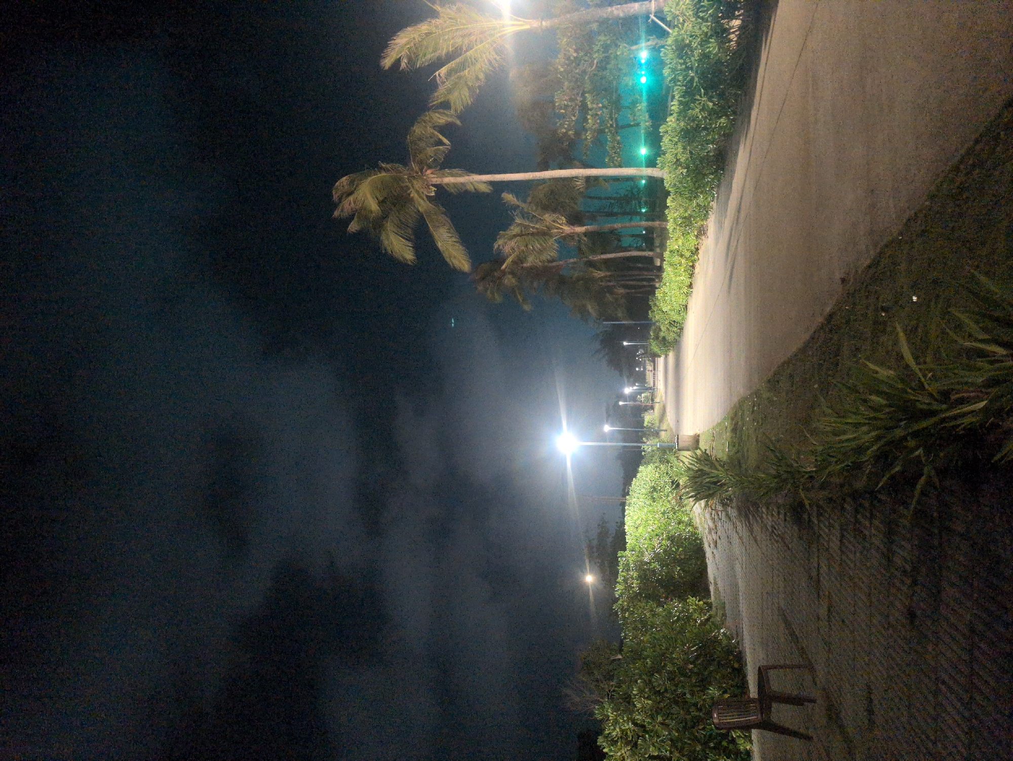 The image depicts a night scene on a pathway in Phú Quốc, Vietnam. A paved walkway, flanked by lush green vegetation and palm trees, stretches into the distance under the illumination of streetlights. The sky is dark, hinting at cloud cover. In the foreground, a simple chair sits on a brick-paved section of the path.