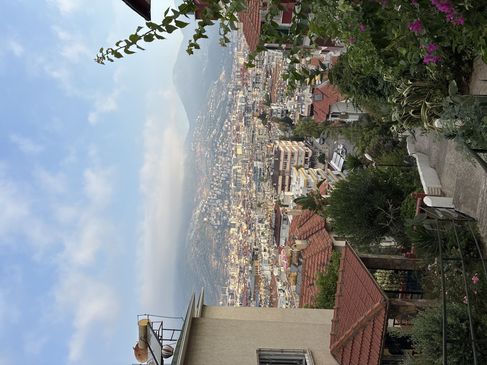 The image depicts a panoramic view of Alanya, Turkey, from an elevated position overlooking the city and the surrounding mountains. In the foreground, a concrete pathway with a black metal railing curves downhill, flanked by lush green foliage and flowering plants, including bright pink bougainvillea. To the left, a light-colored house with a red-tiled roof is partially visible, featuring a solar water heating system on its roof.
The middle ground is dominated by a dense urban landscape filled with multi-story buildings, many with red-tiled roofs. Cars are visible on a street winding through the city. The buildings are clustered closely together, creating a textured urban sprawl. Solar water heaters are a common sight on many rooftops.
In the background, hazy mountains rise, their slopes dotted with more buildings and vegetation. The sky above is a pale blue, with scattered white clouds. The overall impression is a sun-drenched Mediterranean city nestled between the mountains and a distant, unseen coastline, likely captured during the morning or late afternoon given the soft lighting. No people are visible in the image.