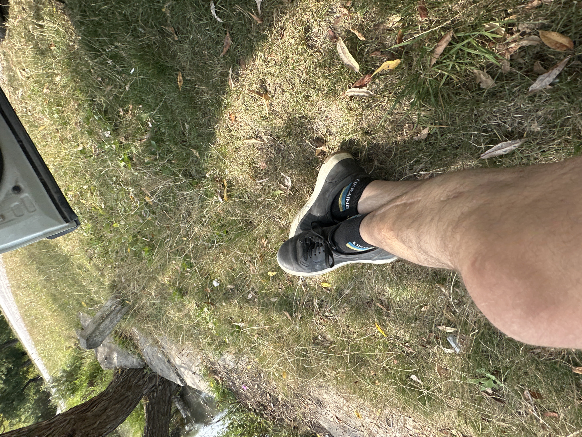 The image shows a person's legs and feet resting on grass outdoors. The person is wearing black sneakers and dark socks with a pattern that includes the word "UKRAINE". The legs are hairy and tanned. The environment appears to be a grassy area with dry grass and some fallen leaves. A tree trunk and some rocks are visible to the left, with a body of water also present in that area. A part of a vehicle door is visible in the upper right corner of the image. The scene is set in Makiv, Ukraine.