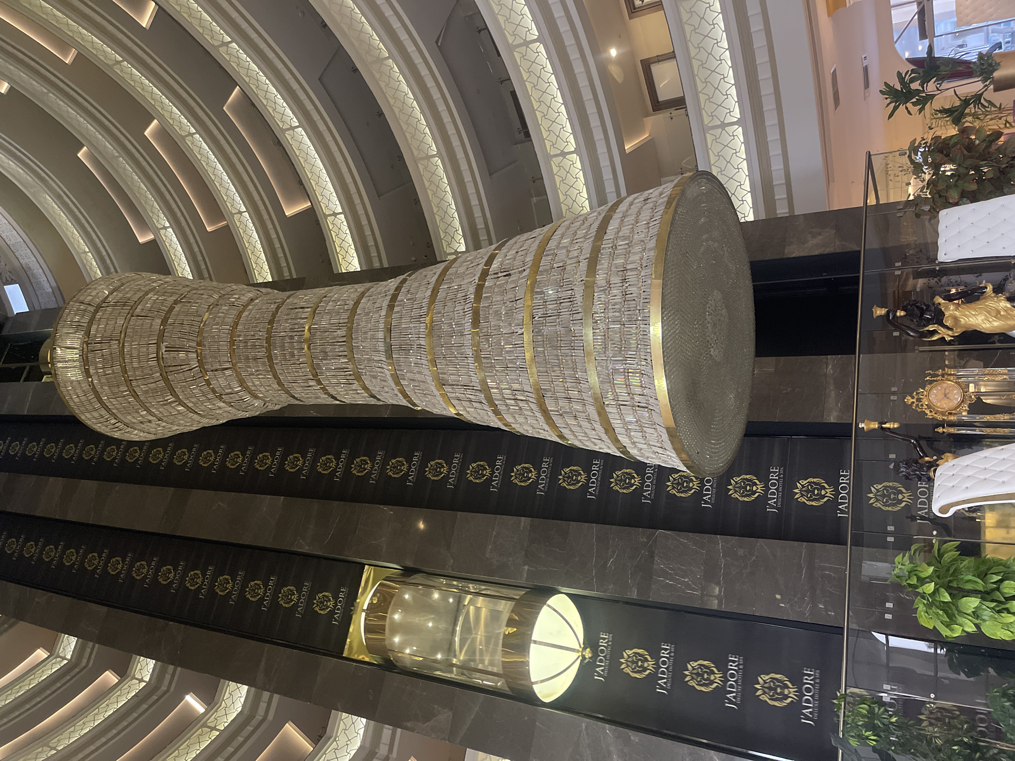 The image displays the grand interior of what appears to be a luxurious hotel atrium, viewed from a low angle looking upwards. A massive, elongated chandelier dominates the central space, constructed from countless shimmering crystal or glass elements arranged in horizontal tiers, accented with gold bands.
The ceiling features multiple ornate, curved arches that ascend vertically, each embedded with patterned lighting that emits a soft glow. Along the dark, polished walls adjacent to the elevators, the brand "J'ADORE DELUXE HOTEL & SPA" is prominently displayed in gold lettering with a distinctive lion's head logo, repeating consistently across the vertical panels.
Two glass panoramic elevators are visible, one at the bottom center of the frame, brightly lit from within, and another partially visible higher up. On the right side of the image, hints of opulent decor include a golden statue, a white quilted bench, an antique-style clock, and some lush greenery. The scene is static, devoid of people, emphasizing the architectural grandeur and luxurious ambiance of the space.