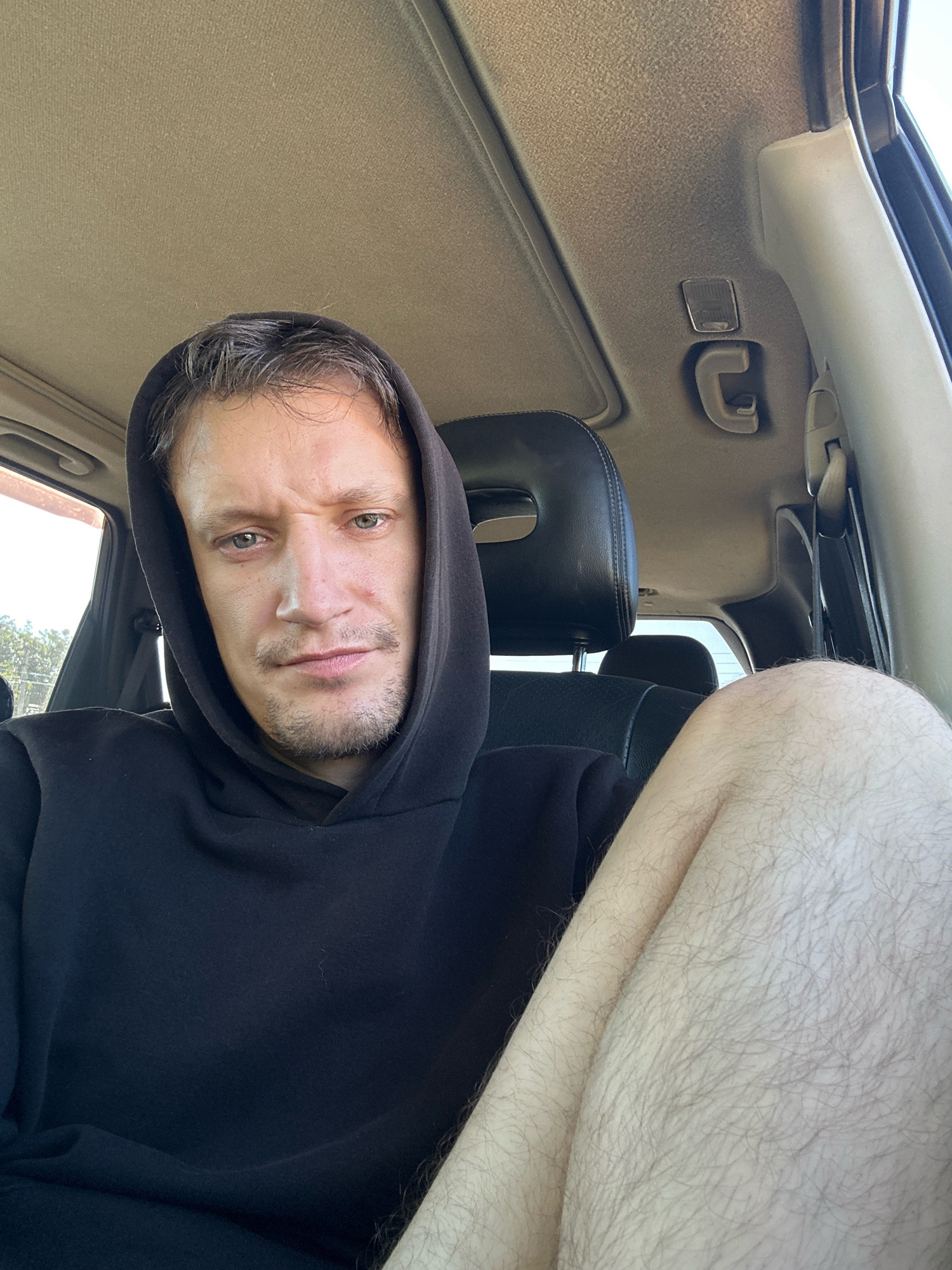 In Makiv, Ukraine, a man with short brown hair and stubble is seated in the passenger seat of a car. He is wearing a black hooded sweatshirt and his bare leg is visible. The interior of the car is visible, including the headrest and ceiling. The man is looking directly at the camera.