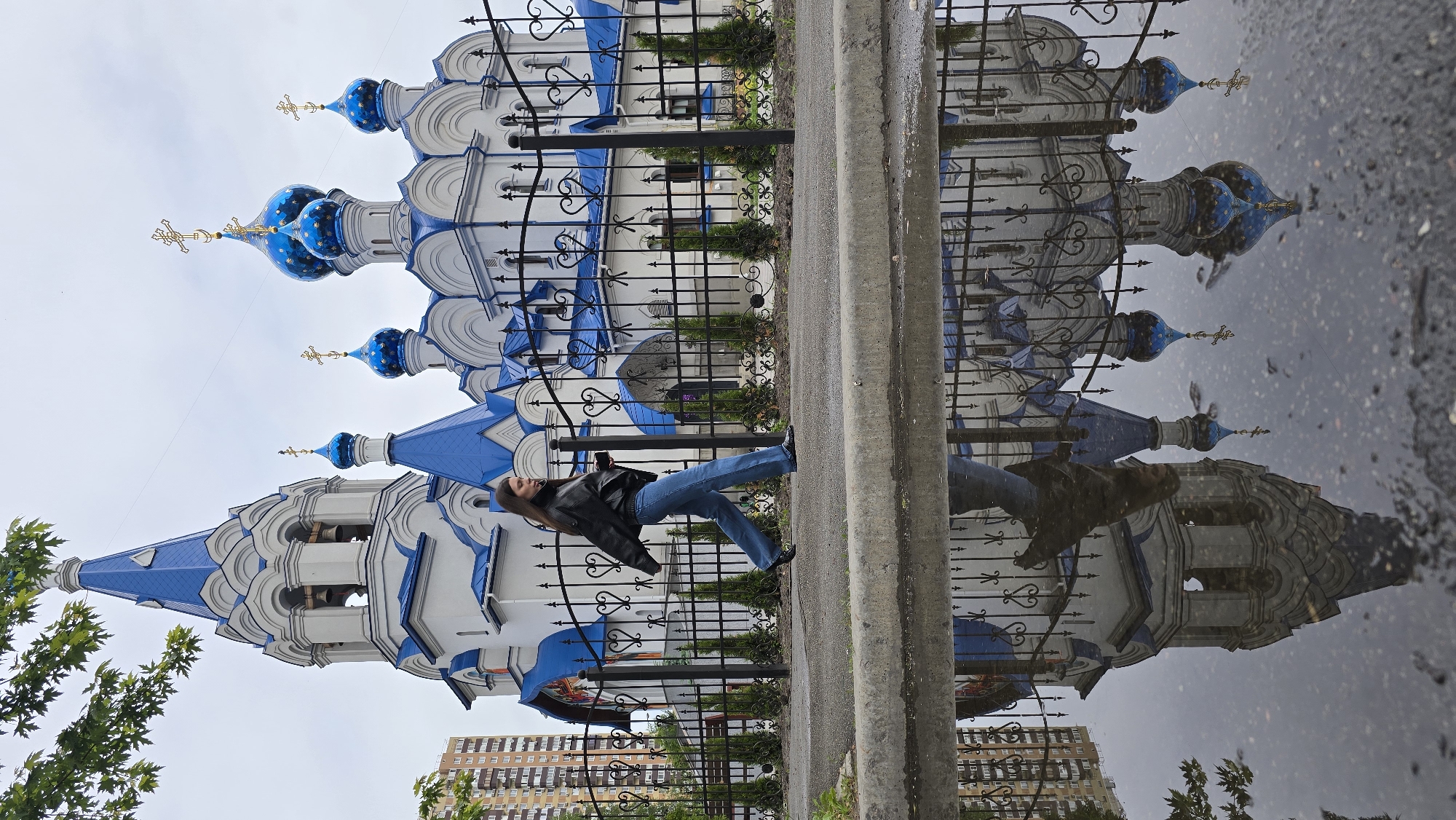 An upright (after 90-degree counter-clockwise rotation) full-length shot captures an ornate Orthodox church, a woman, and their reflections in a large puddle in Kyiv, Ukraine. The setting appears to be an urban street or sidewalk following recent rainfall.
The prominent subject is a beautiful Orthodox church, distinguished by its white walls and multiple bright blue onion domes, each topped with a golden cross. The church features intricate architectural details, including rounded arches and decorative trim. A tall bell tower with a blue roof and smaller domes is visible on the left side of the church.
In the foreground, a woman stands near a decorative black wrought-iron fence. She is wearing a black jacket and blue jeans, with dark hair cascading down her back. She is looking towards the church, seemingly observing its architecture or posing for a picture.
A significant element of the image is the large puddle that occupies the lower half of the frame, perfectly mirroring the church, the fence, and the woman. The reflection is clear, though slightly distorted by ripples or debris on the water's surface, lending an artistic quality to the scene. Distant multi-story residential buildings and green foliage are also partially visible in the background, both directly and in the reflection.
The sky is overcast, providing soft, diffused lighting that highlights the vibrant blues and whites of the church. The presence of the puddle strongly suggests a recent rain shower. There is no visible text in the image. The scene captures a moment of serene contemplation against a backdrop of striking religious architecture, enhanced by the natural mirror of the rainwater.