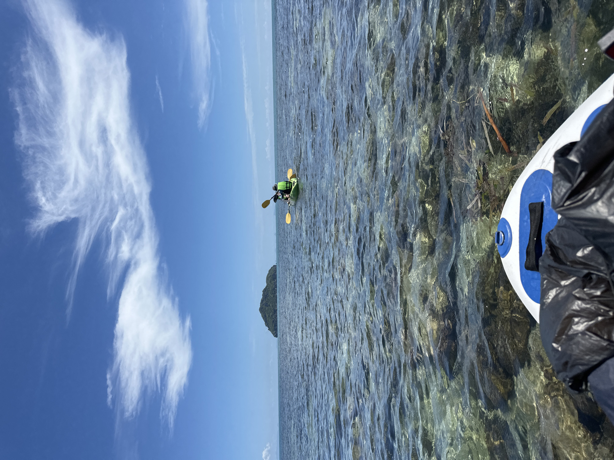 This is a landscape photo taken from a first-person perspective, likely from a paddleboard, looking out at a clear ocean with visible coral and seaweed on the seafloor. In the middle distance, two individuals are kayaking in a green kayak, both wearing life vests and holding paddles. They are paddling towards a small, tree-covered island on the horizon. The sky above is a vibrant blue with wispy white clouds. The scene depicts a sunny day on the water, suggesting a recreational activity in a tropical location. While the specific location within ตำบลเกาะพะงัน, Thailand, cannot be precisely identified from the image alone, the visual elements are consistent with the coastal scenery found in that region.