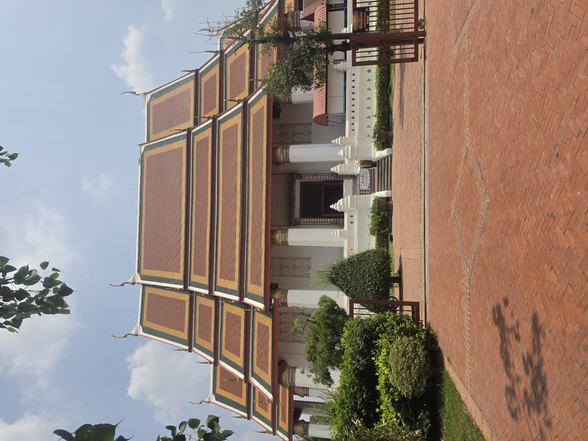 A building with a tiled multi-tiered roof stands in Phra Nakhon Si Ayutthaya City Municipality, Thailand. The roof tiles are rectangular and maroon, trimmed with yellow and green. The building has a white facade with columns. A red brick walkway leads alongside the building. Green shrubbery grows in front of the building. A tree is visible on top of the roof. The sky is light blue with scattered clouds.
