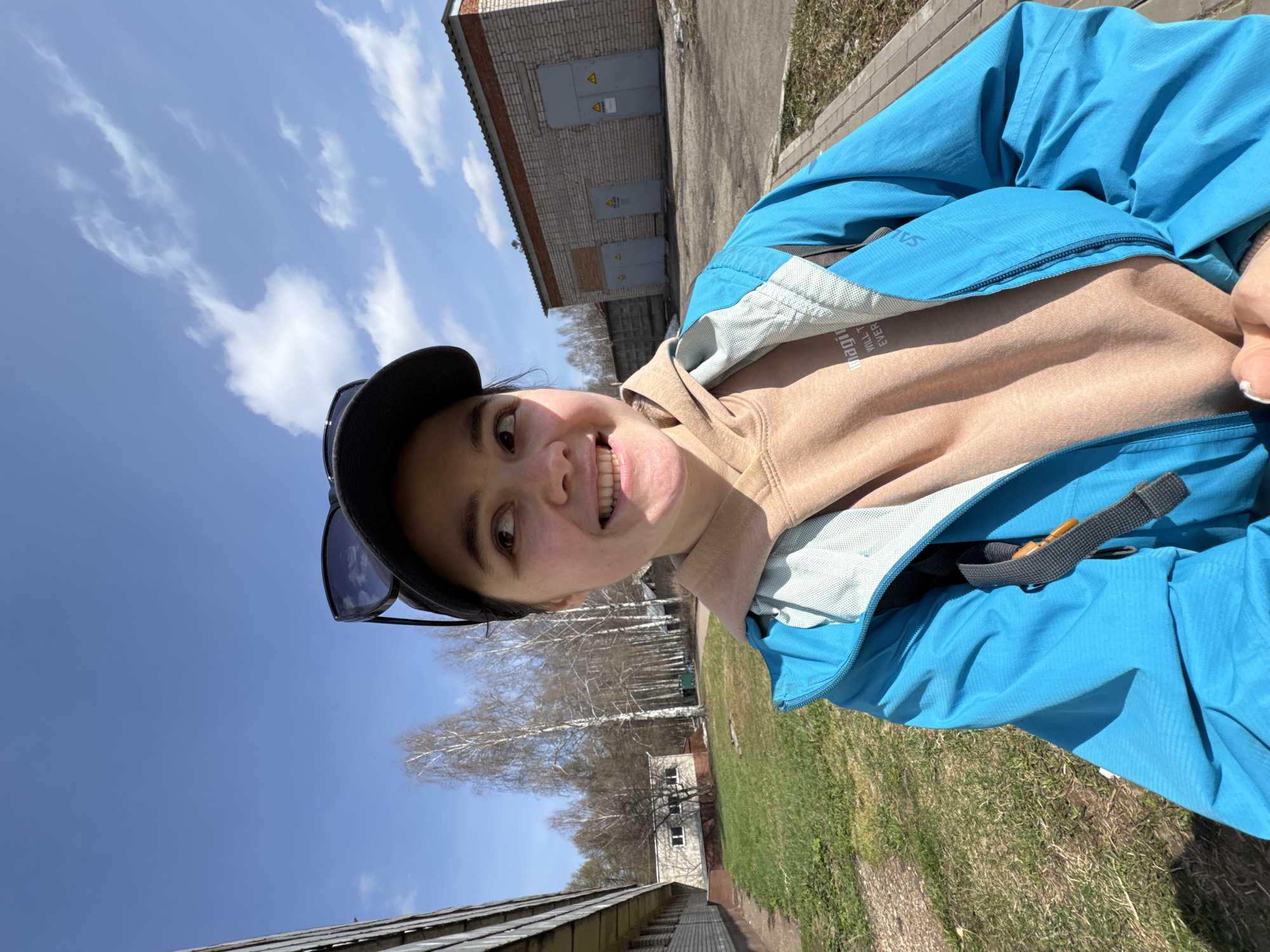 A selfie captured outdoors shows a woman smiling at the camera, wearing a blue jacket, a tan hoodie, and a black baseball cap with sunglasses resting on top. The background includes a clear blue sky with scattered clouds, suggesting a sunny day. Behind her, a utility building made of light-colored bricks with visible electrical hazard signs can be seen. Additionally, there is a grassy area and a line of birch trees in the distance. The photo appears to have been taken near an electrical substation, which could be located on the outskirts of Izhevsk, Russia, considering the presence of such infrastructure. The image orientation suggests the woman is holding the camera above her head, angling it slightly downward.